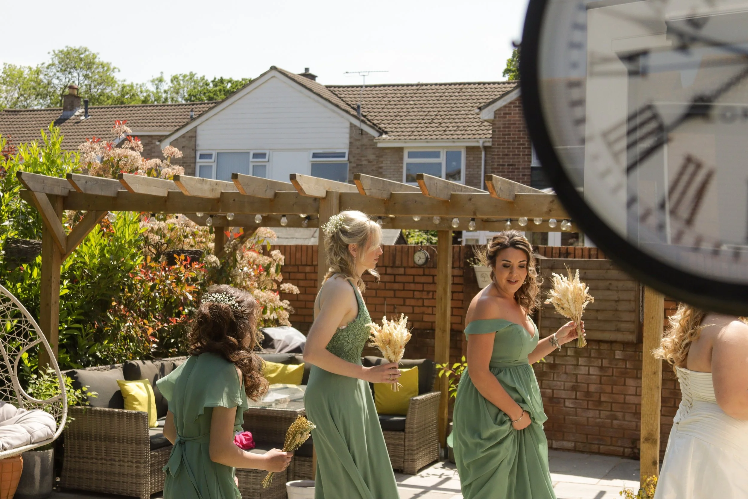 Women and a girl in green dresses holding bouquets, participating in a wedding ceremony outdoors on a sunny day, with a brick wall, wooden pergola, garden furniture, and houses in the background.