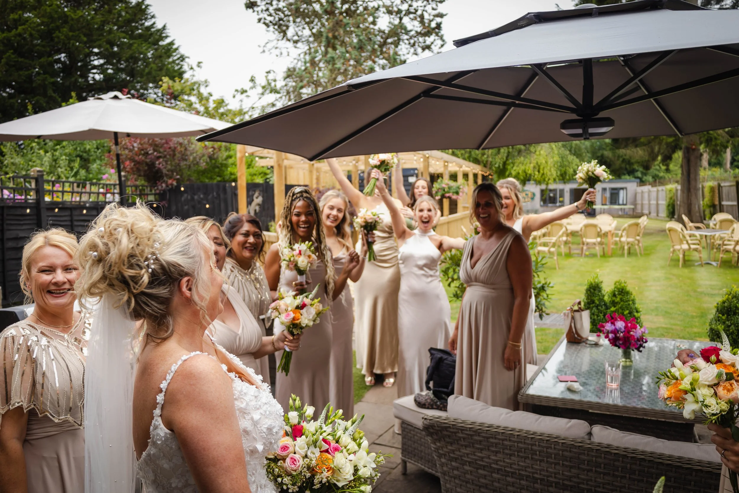 Group of women at a wedding reception outdoors, some holding bouquets, smiling and celebrating under umbrellas in a garden setting.
