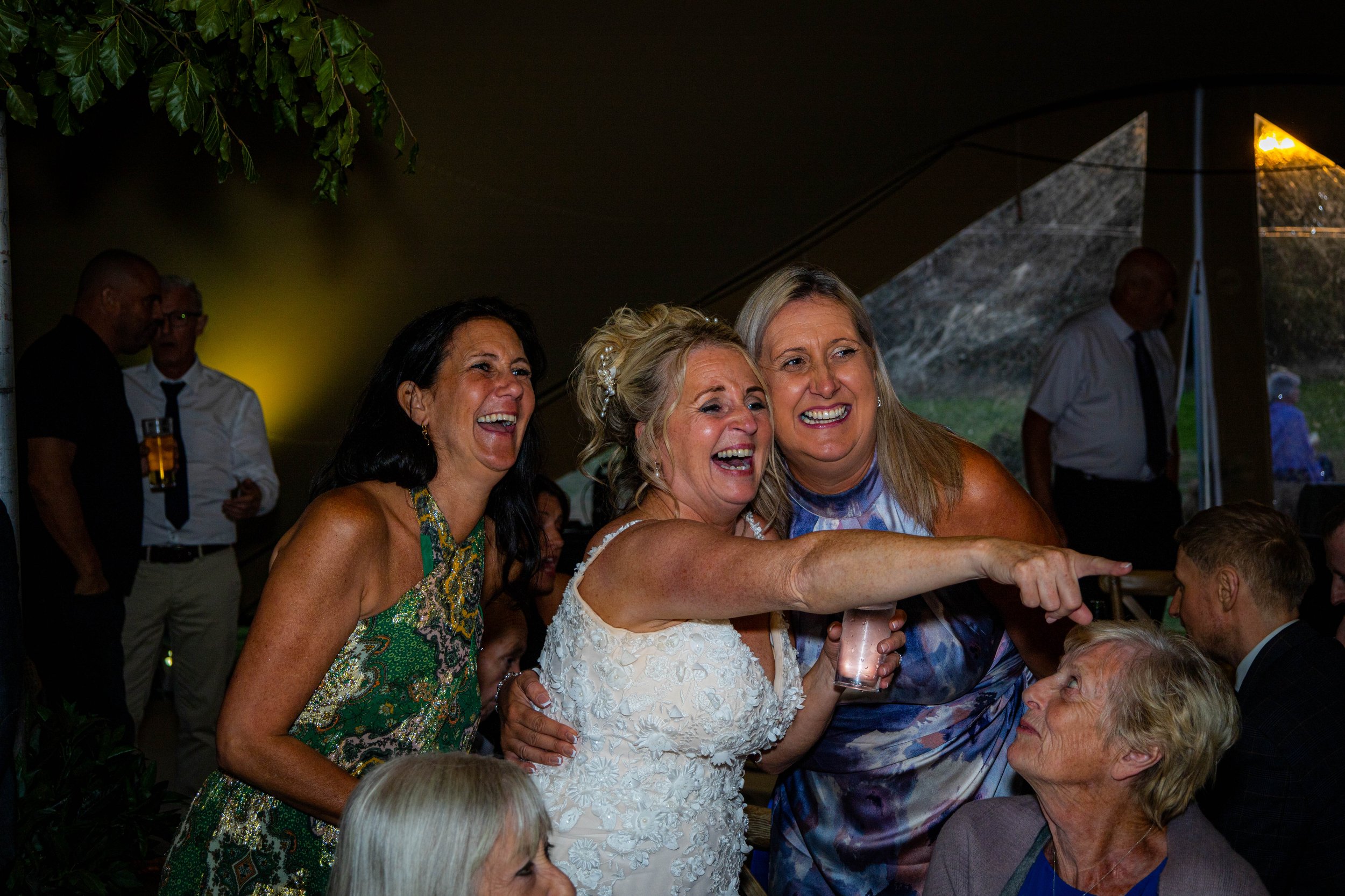 Group of women smiling and laughing at a celebration, possibly a wedding reception, with some women pointing and others holding drinks.