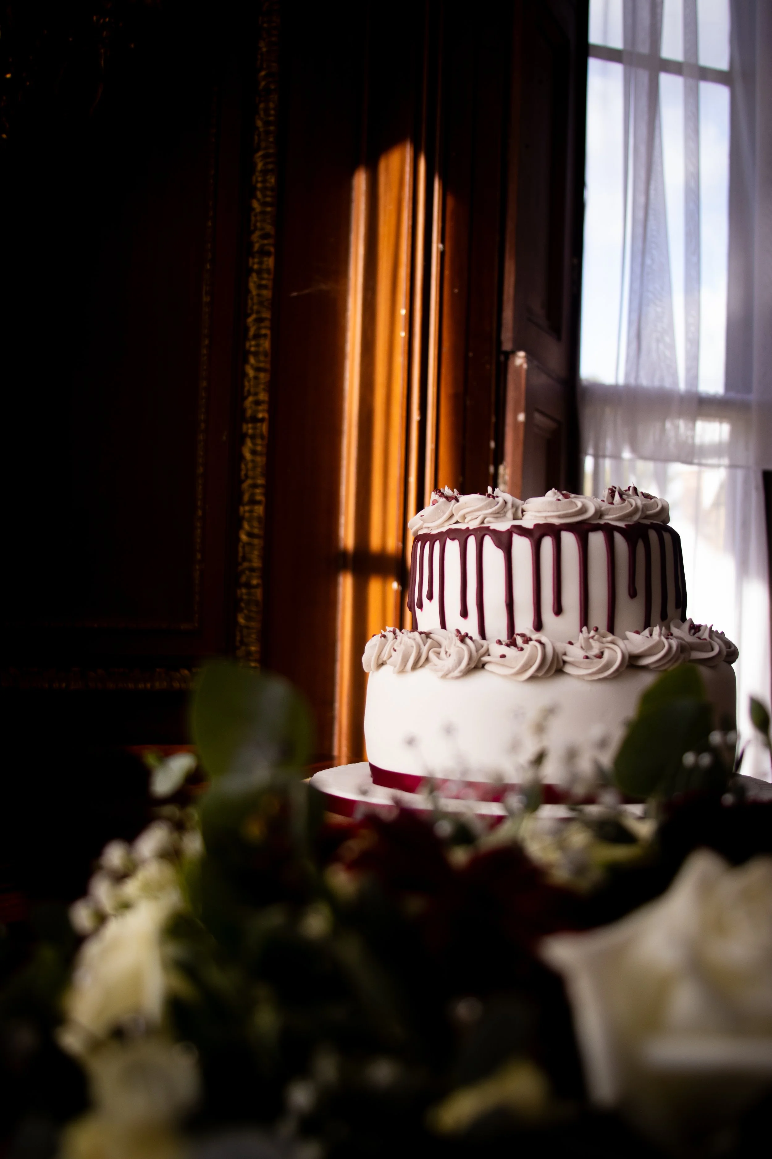 Two-tiered wedding cake decorated with white icing, pink drips, and pink frosting swirls, with greenery and white flowers in the foreground, set in a room with wooden paneling and a window with sheer curtains.