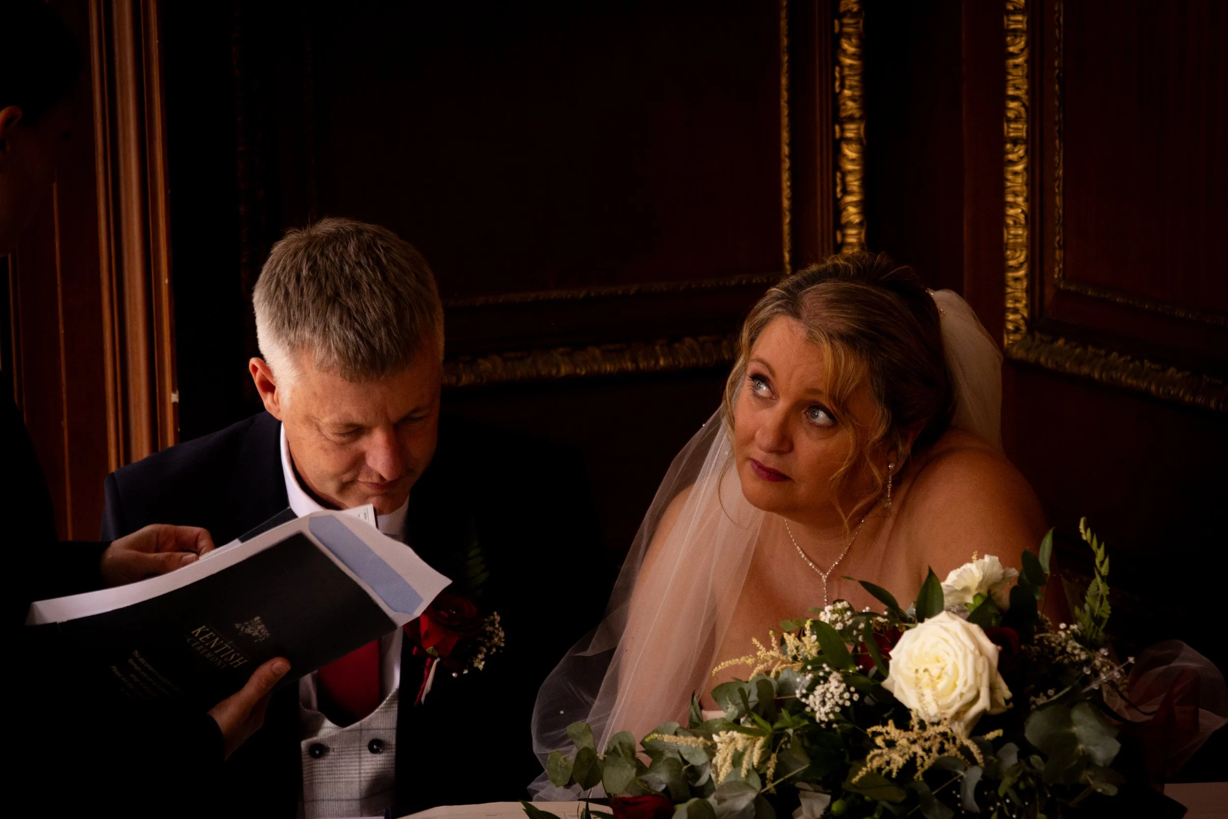 A bride in a wedding dress and veil with a bouquet of white roses and greenery, sitting at a table looking up thoughtfully, during her wedding ceremony with a man in a tuxedo reading from a book.