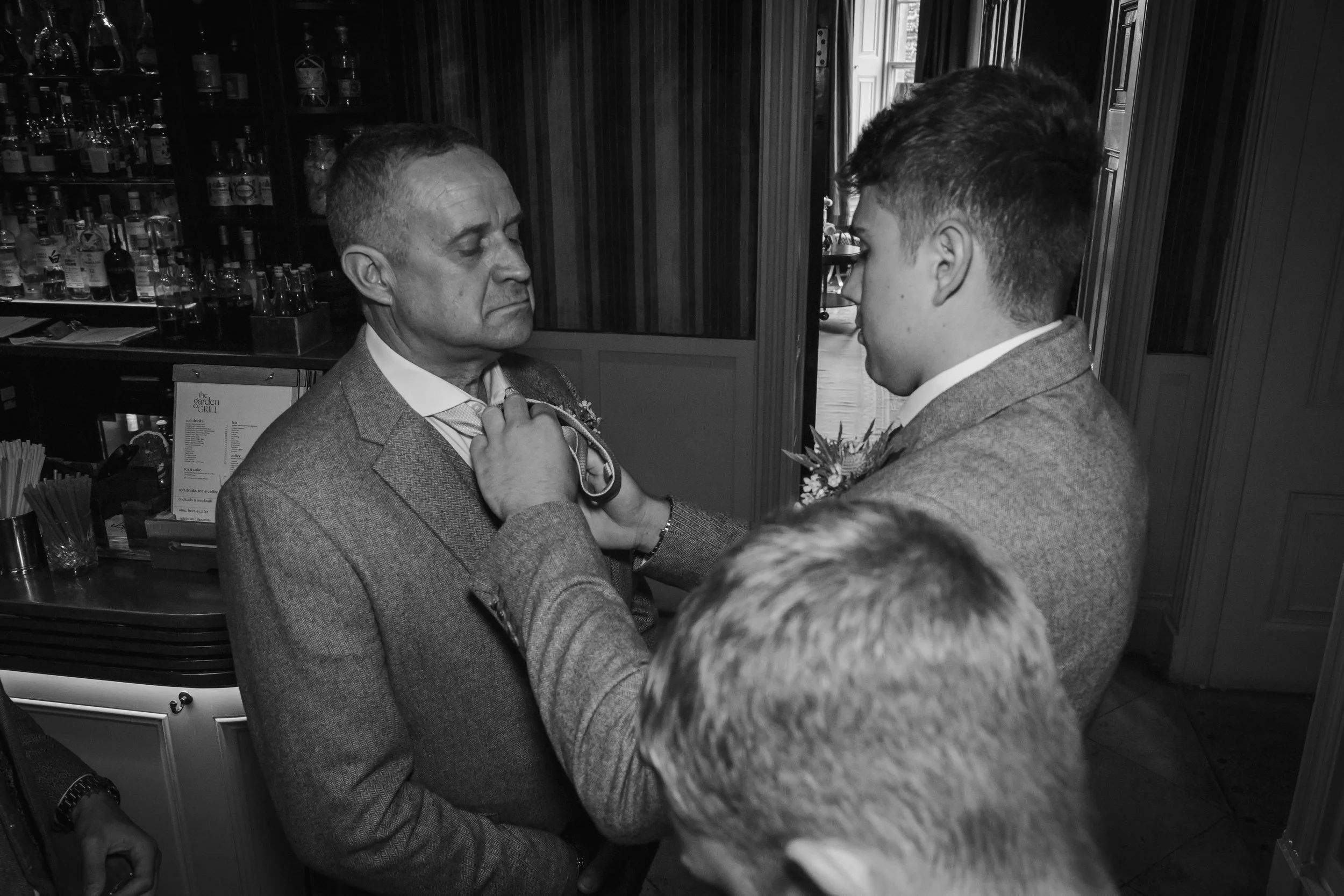 A young man adjusting a bow tie on an older man in a suit inside a bar or restaurant.