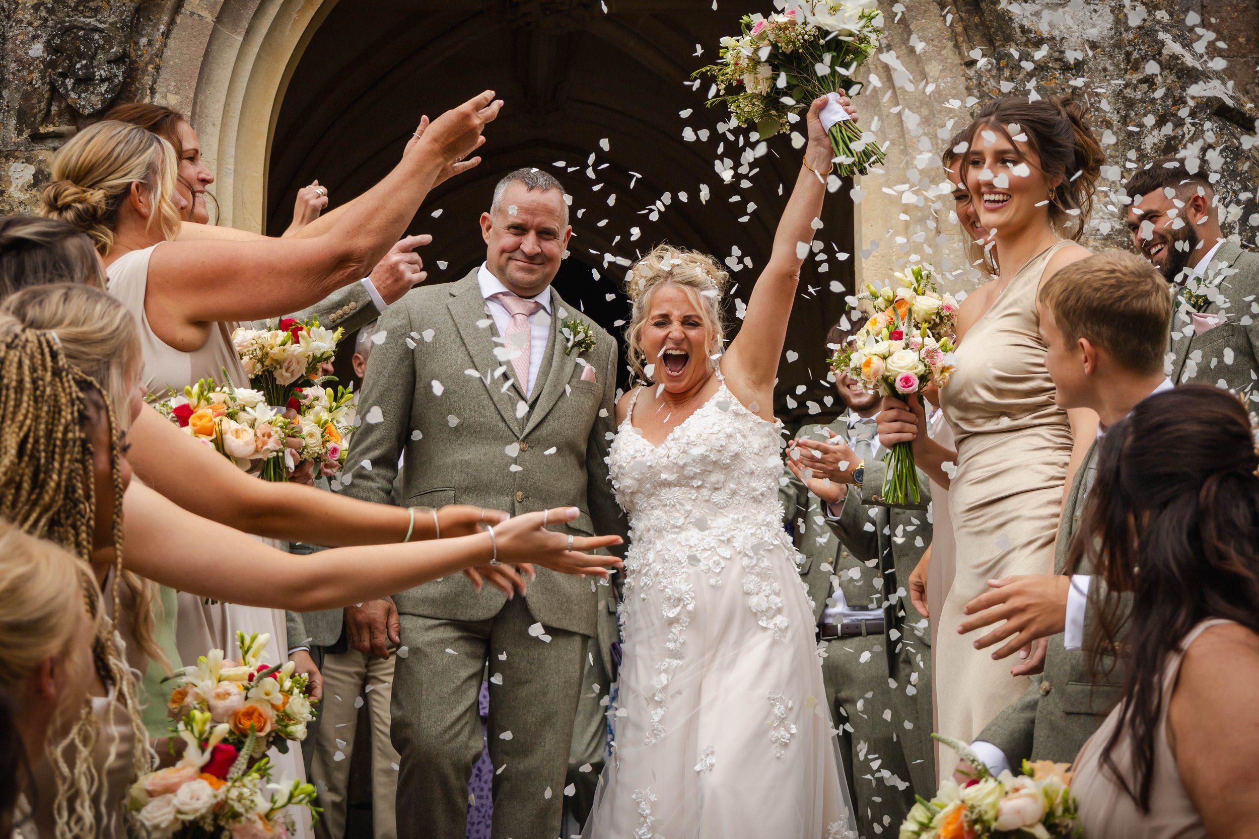 A joyful bride in a white wedding dress, throwing flower petals, surrounded by smiling friends and family during a wedding celebration outside a historic building.