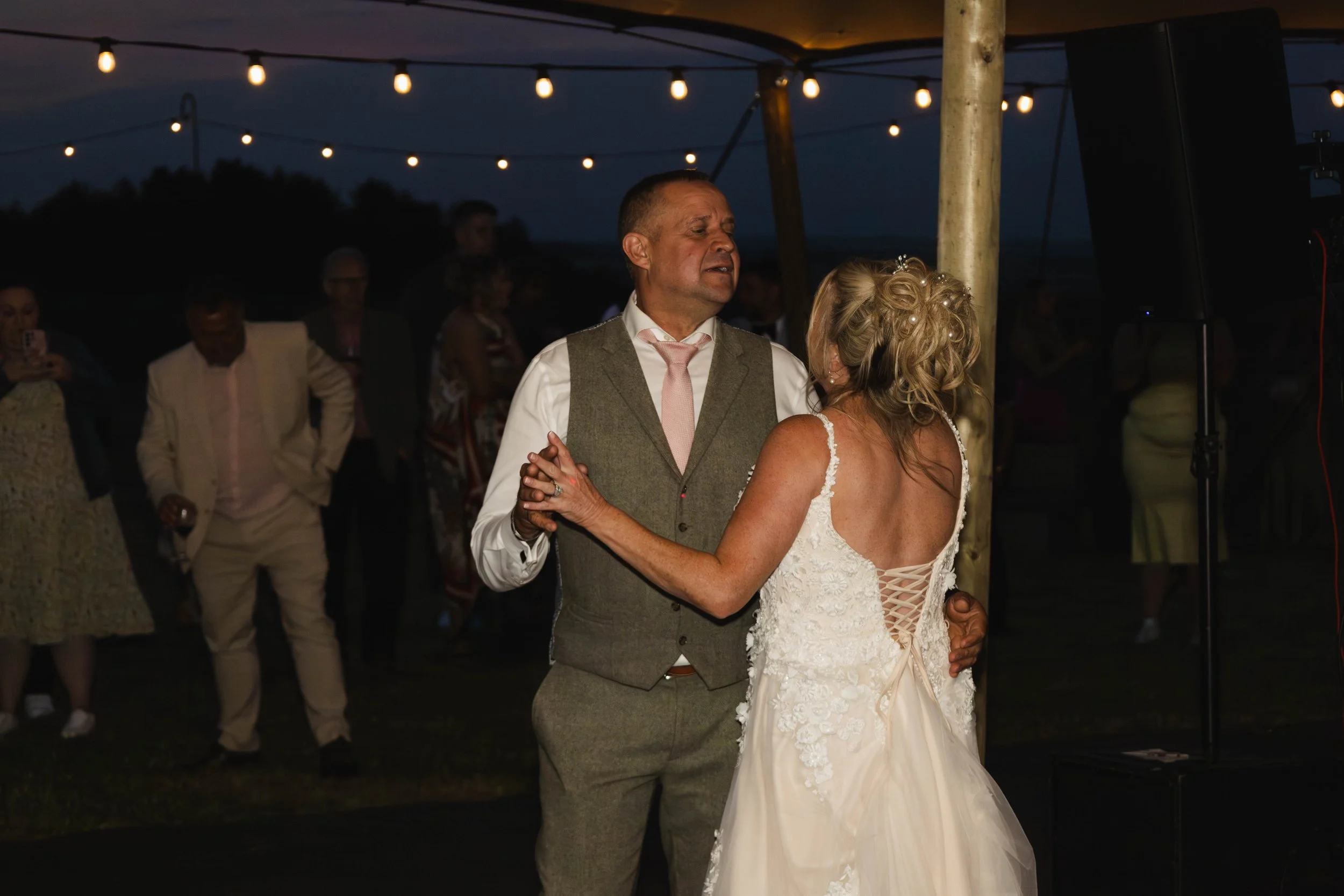 A bride and groom dance at their wedding reception during nighttime outdoors, with string lights overhead and guests in the background.