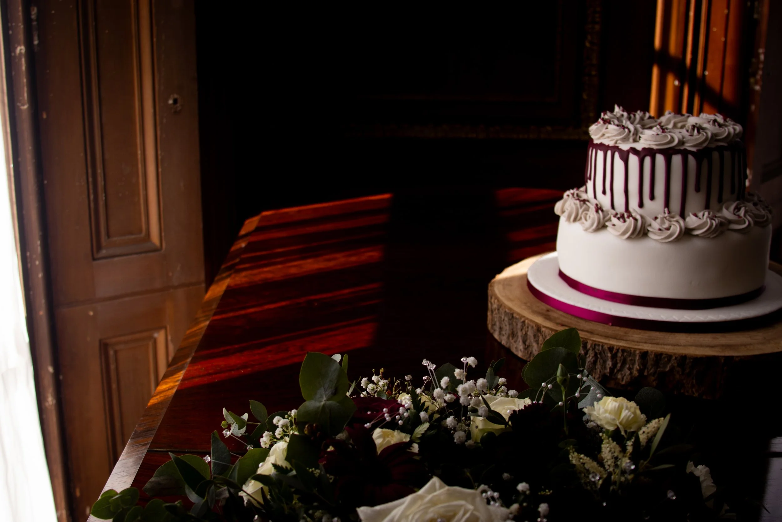 A white, two-tiered wedding cake with purple drips and purple ribbon at the base, topped with white frosting rosettes, sitting on a wooden slab; in front, a bouquet of white flowers and green leaves.