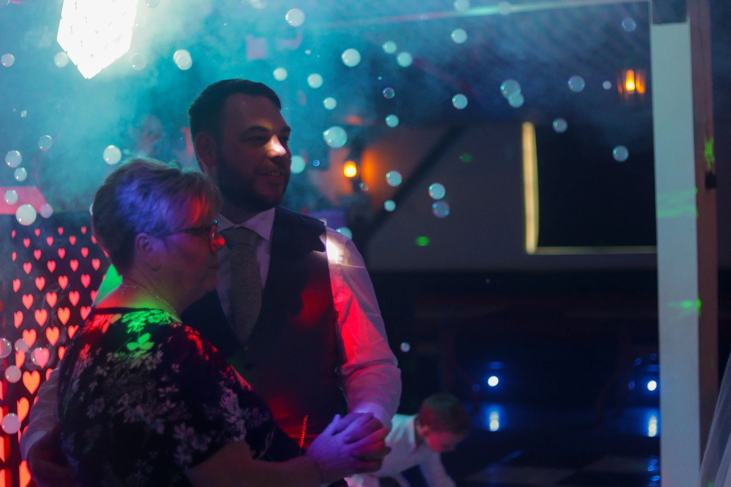 Two people, an older woman and a man, dance in a dimly lit, colorful party or wedding reception with heart patterns and bubbles in the background.