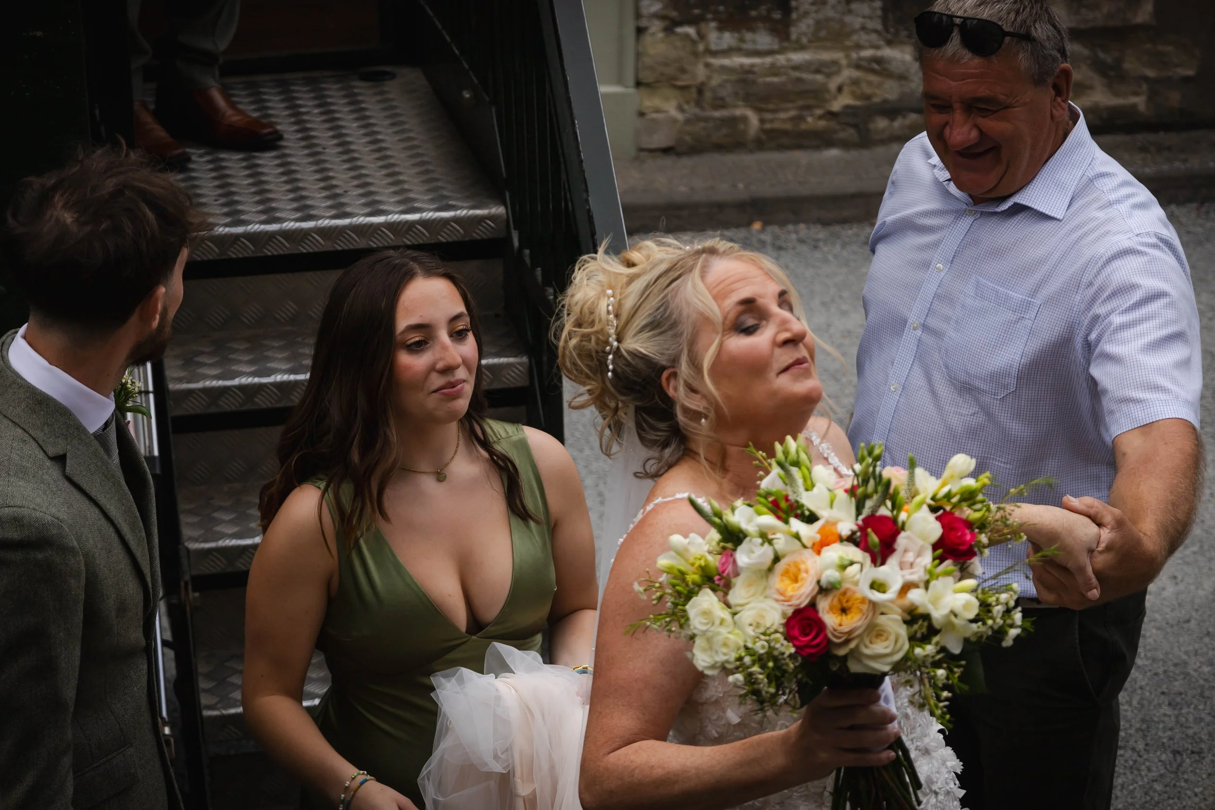 A bride with a bouquet of colorful flowers is being assisted by an older man, possibly her father, at her wedding. Two younger people, a man and a woman, are observing nearby, standing outside near stairs and brick wall.