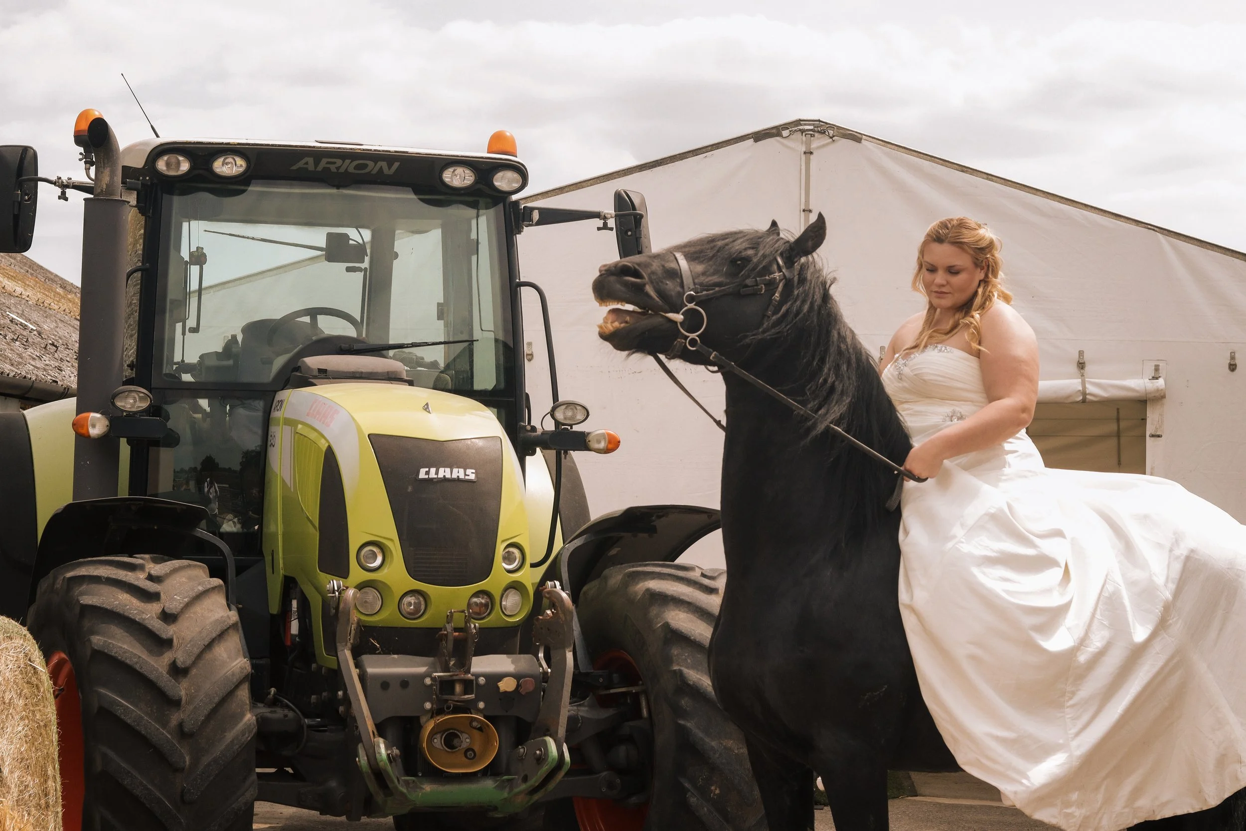 A woman in a white wedding dress riding a black horse in front of a green tractor.