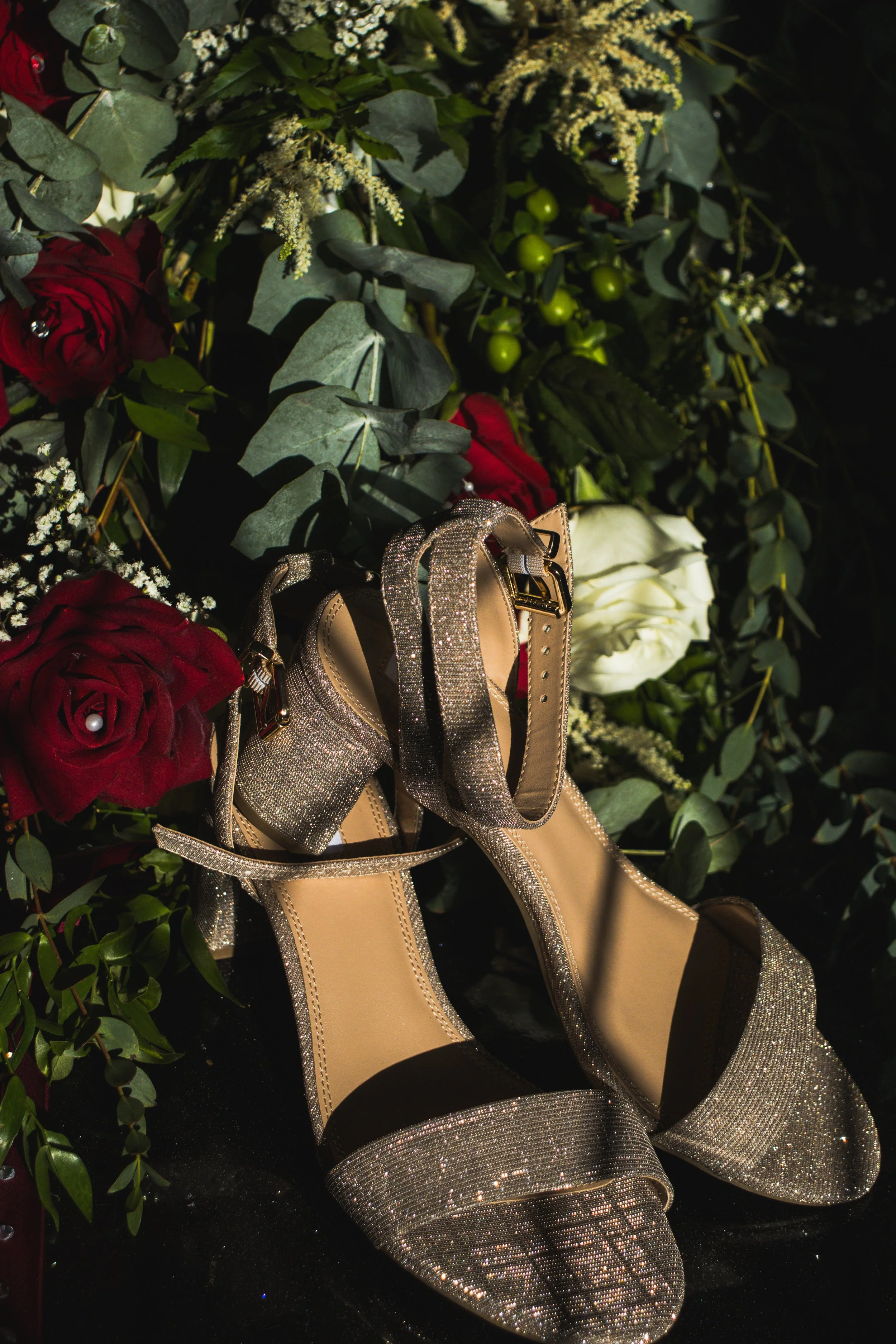 A pair of sparkly high-heeled shoes with ankle straps are placed among a bouquet of red and white roses and greenery.