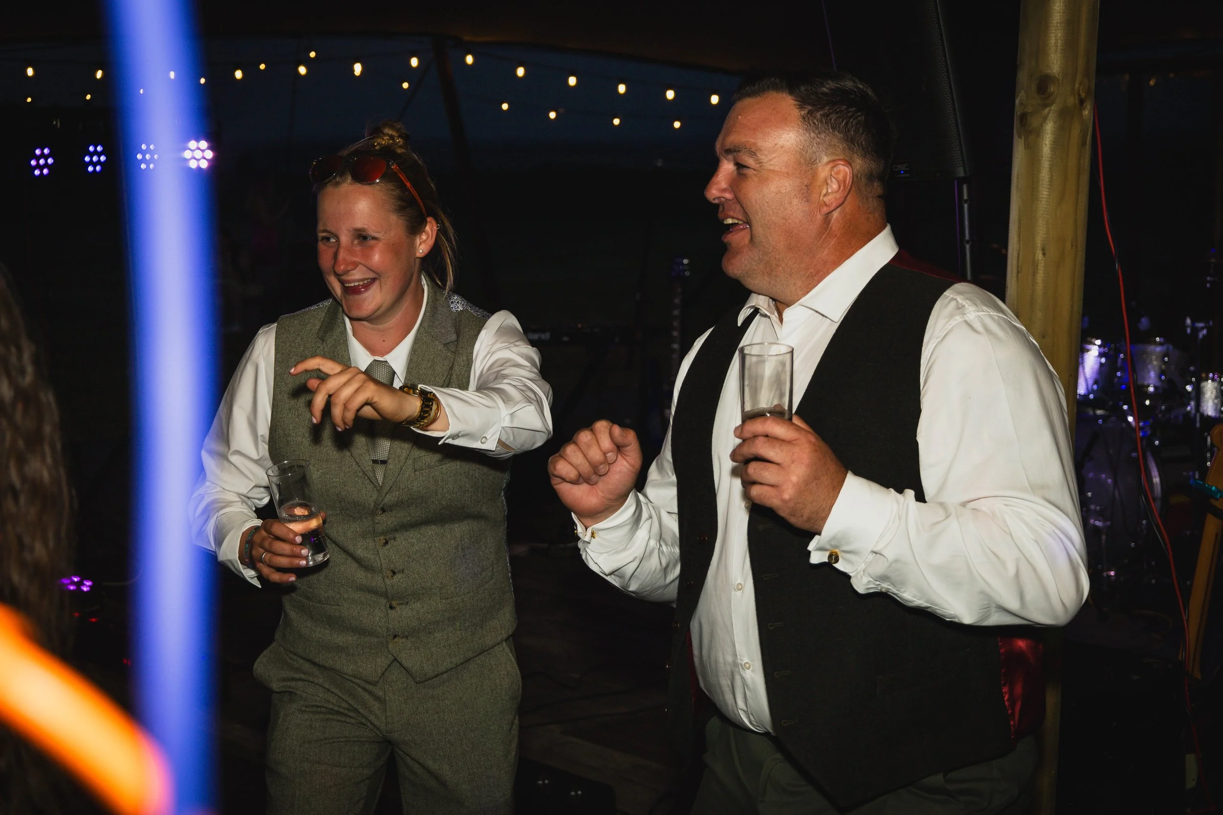 Two people, a woman and a man, dancing and smiling at a party or celebration, holding glasses of drinks, in a dimly lit setting with string lights overhead.