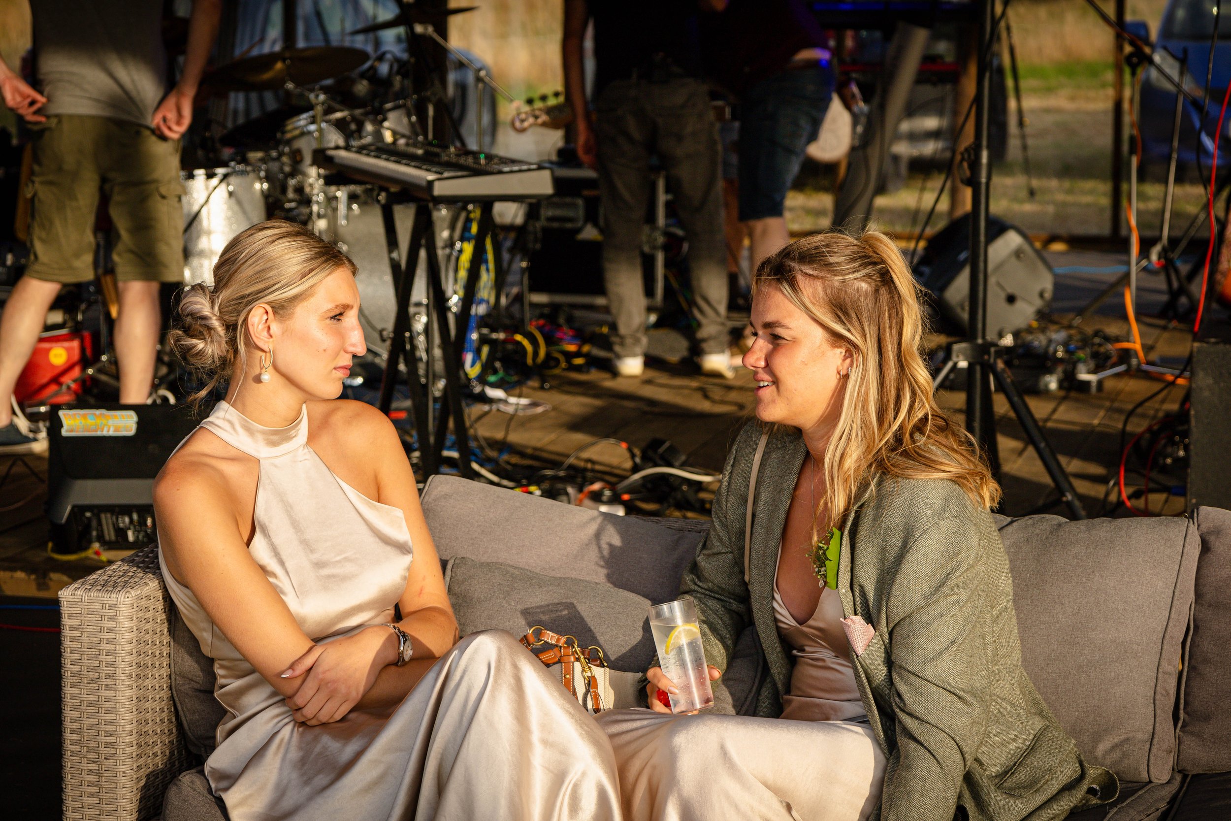 Two women sitting on a couch, having a conversation at an event with electronic equipment and people in the background.