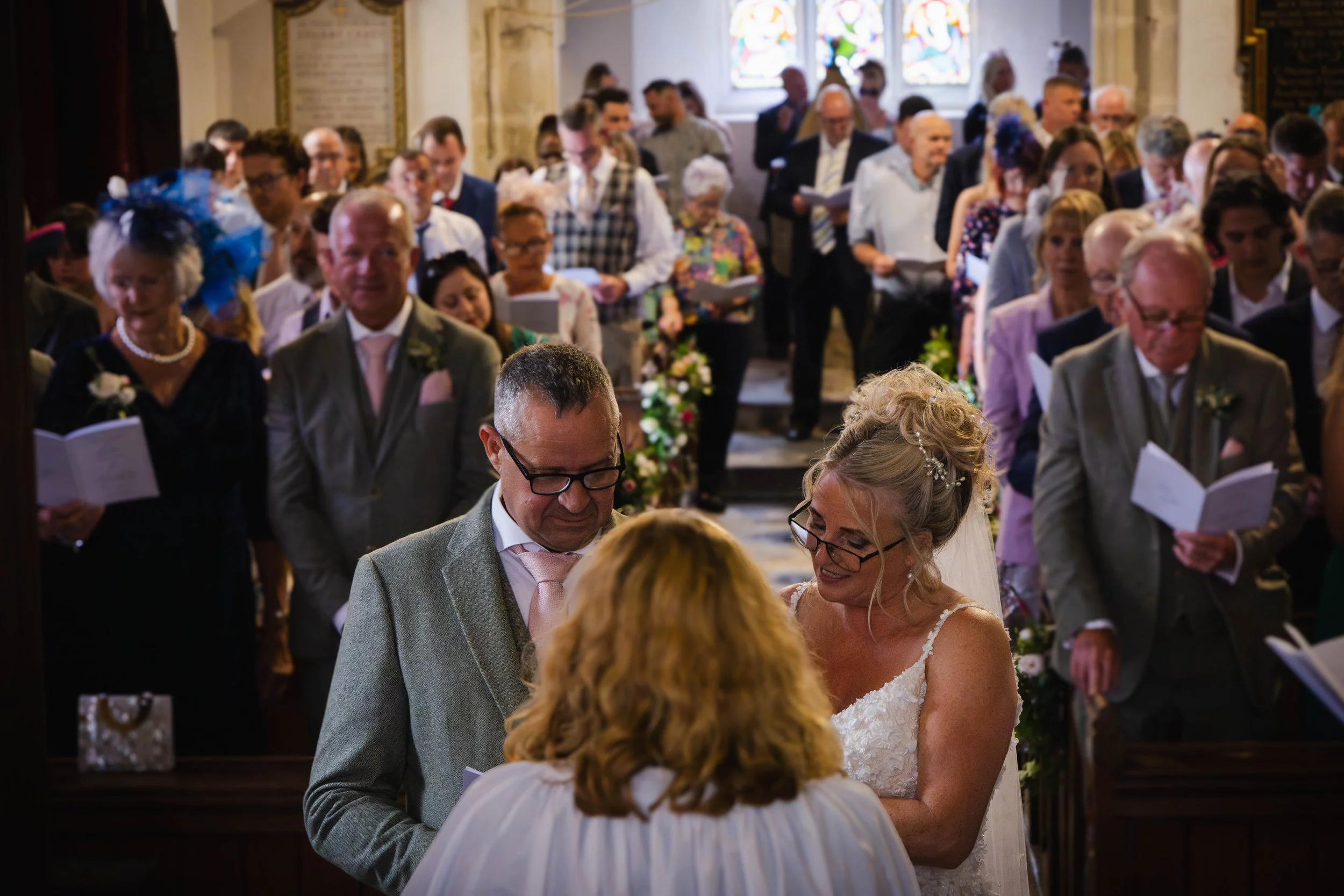 A wedding ceremony inside a church, with a bride and groom standing before an officiant, surrounded by guests seated and standing, holding programs, with stained glass windows in the background.