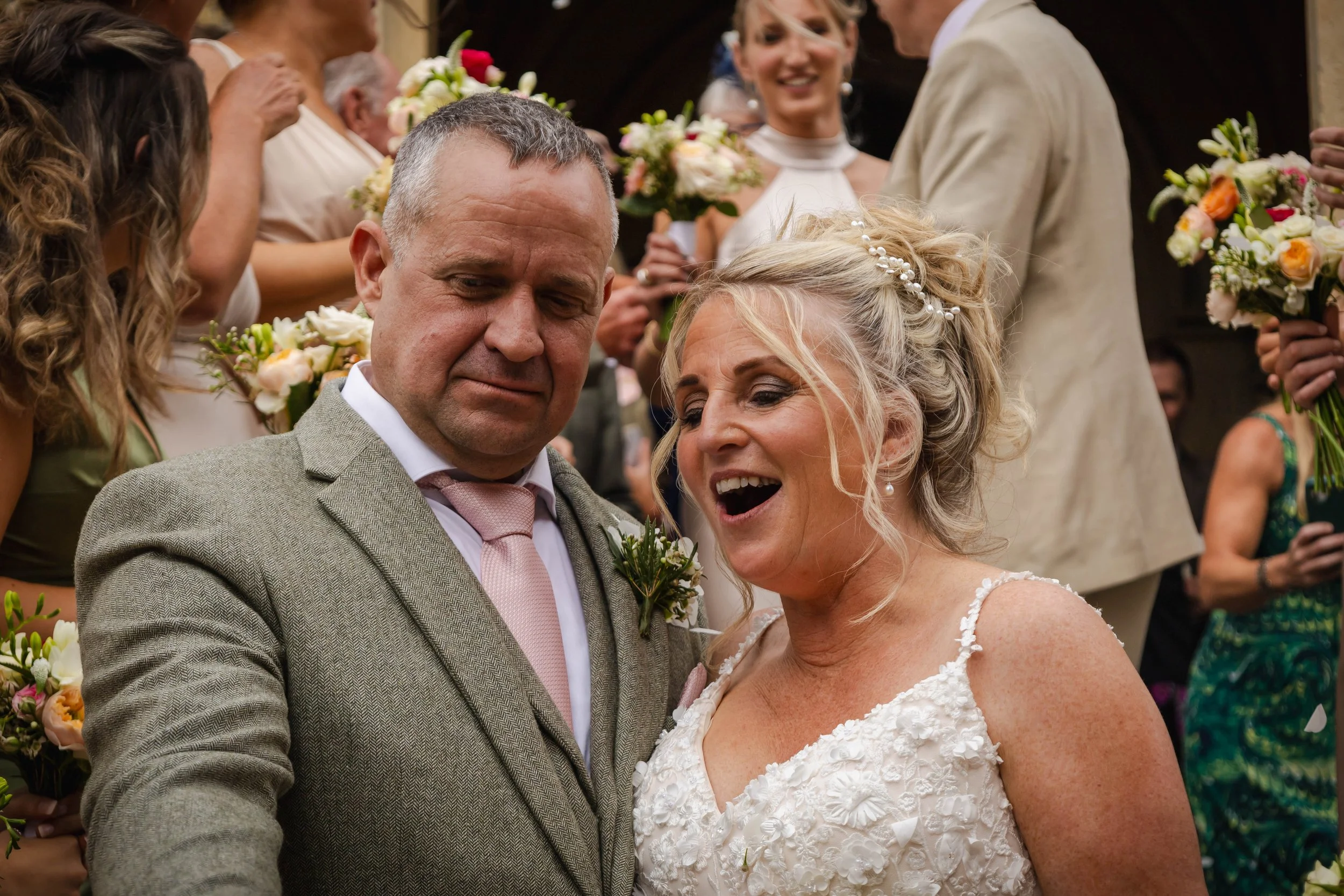 A woman in a wedding dress with a floral appliqué design and a man in a suit with a pink tie, surrounded by bridesmaids holding bouquets, at a wedding celebration.