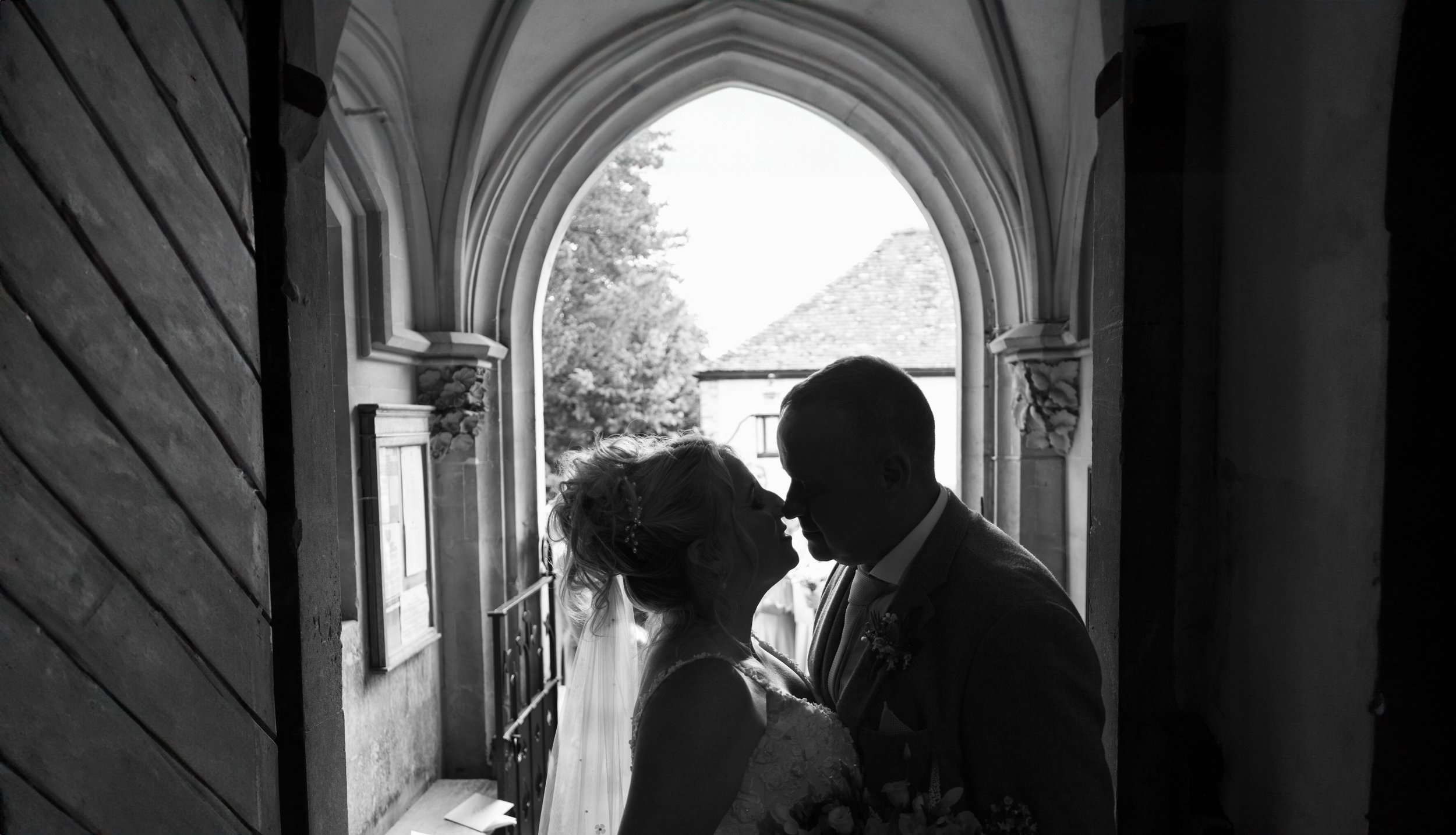 Silhouetted bride and groom sharing a kiss in a church doorway with an arched window in the background.