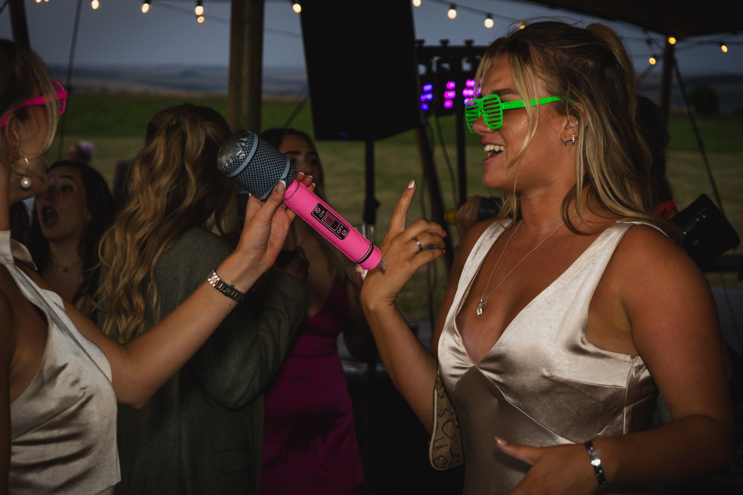 woman at a party wearing green shutter shades, smiling and talking into a pink microphone held by another woman with pink sunglasses, other women in the background under a tent with string lights, outdoor setting during evening.