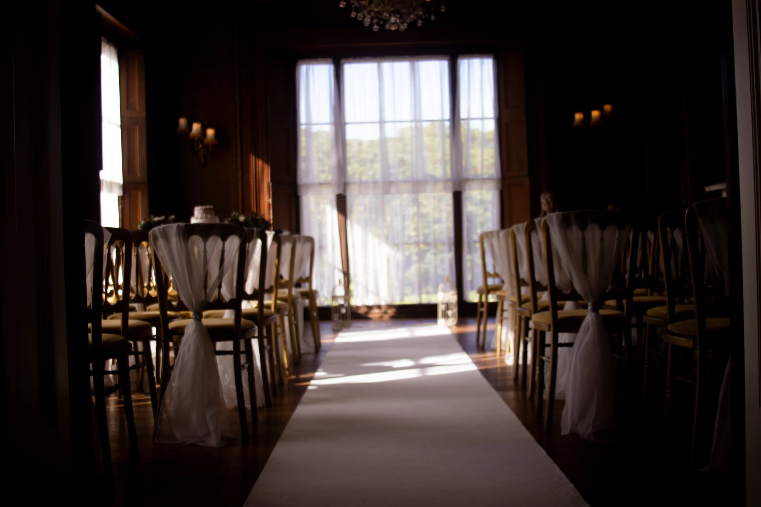 A dimly lit room with wooden walls and large windows covered with sheer curtains, decorated for a wedding or event, with a white aisle runner and chairs draped with white fabric.