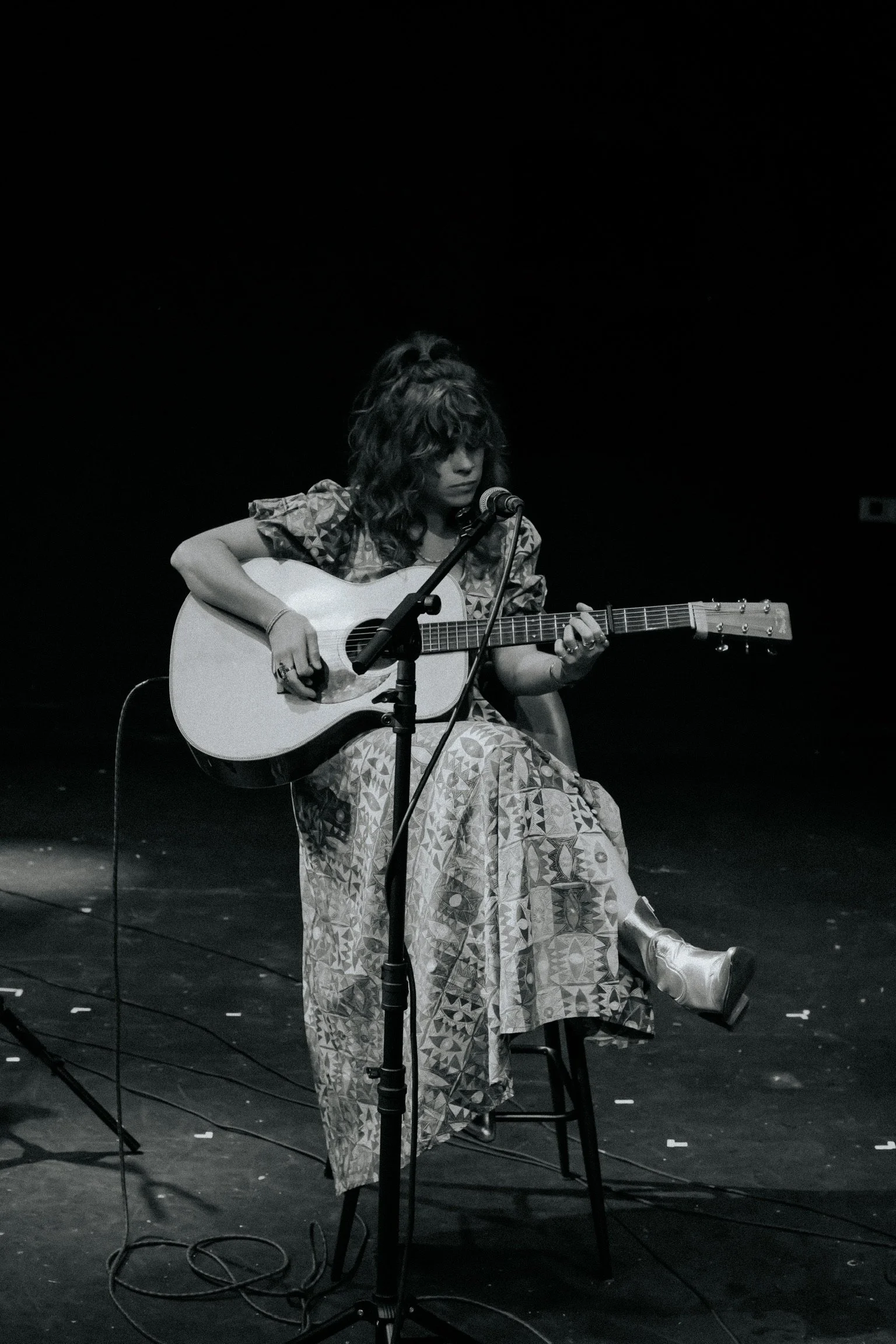 A woman sitting on a chair playing an acoustic guitar on a dark stage with a microphone in front of her, in black and white.