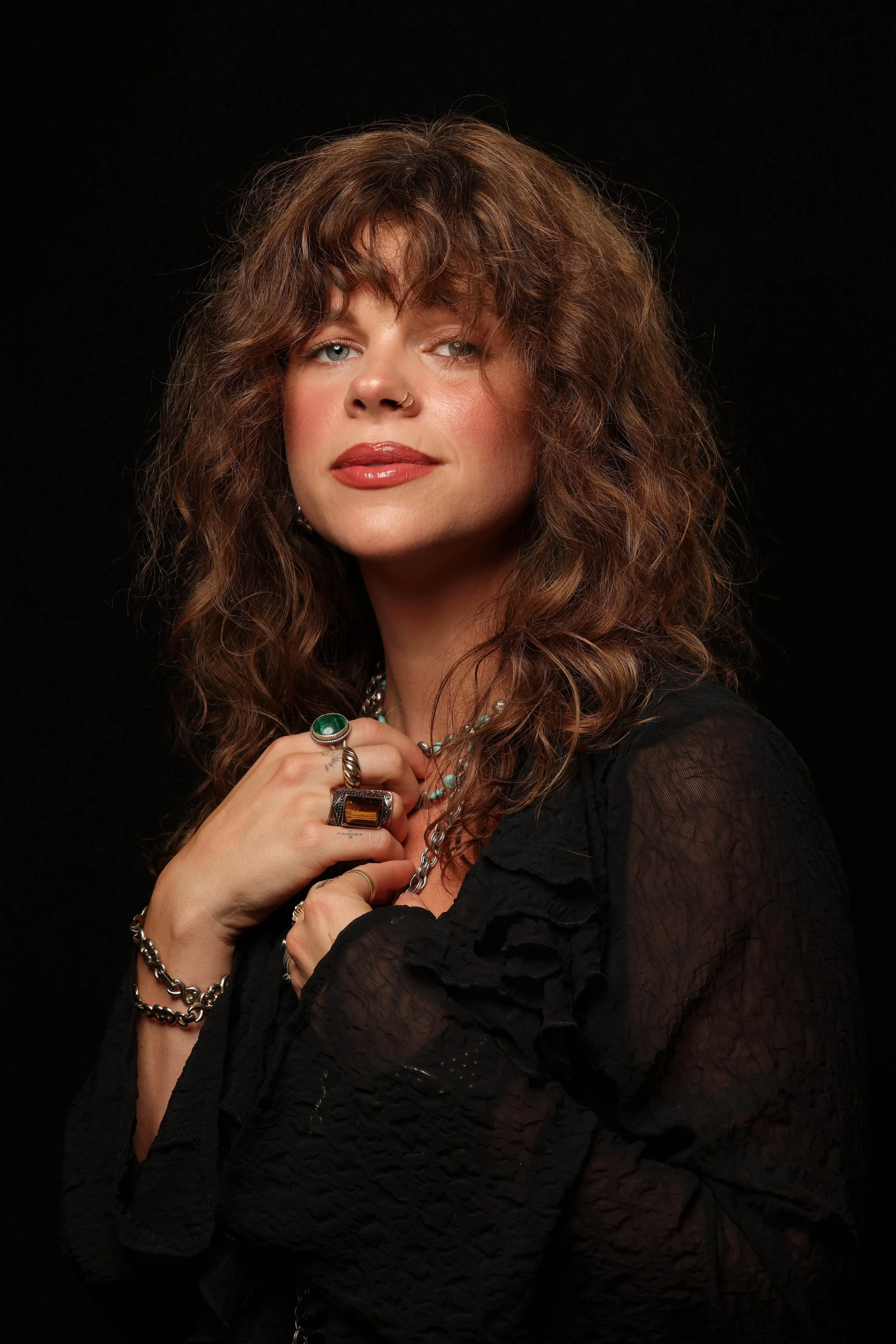 A woman with curly brown hair and blue eyes wearing a black lace top, adorned with rings, necklaces, and bracelets, posing against a black background.