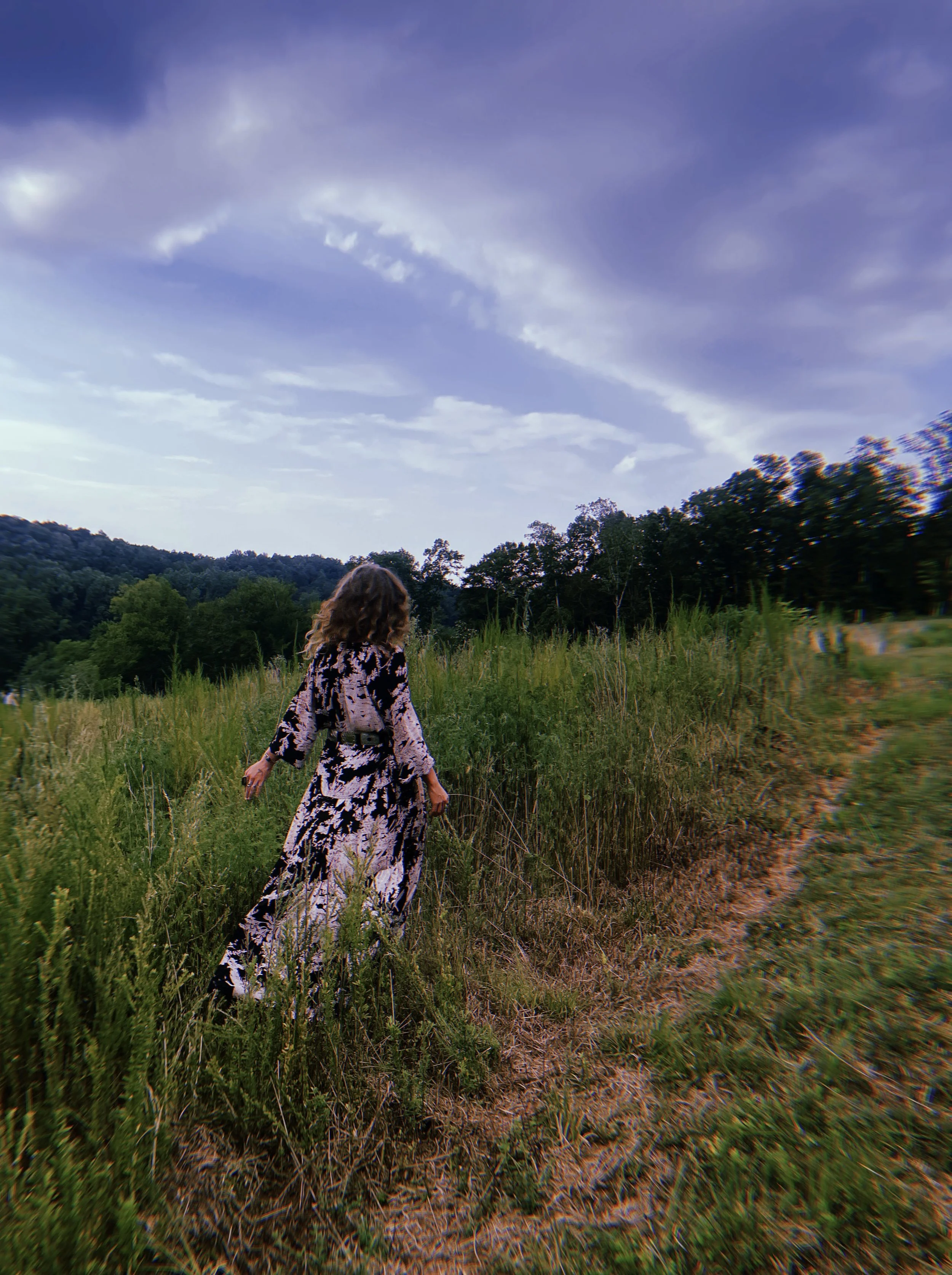 Woman in a black and white patterned dress walking through a grassy field with trees and hills in the background, under a partly cloudy sky.