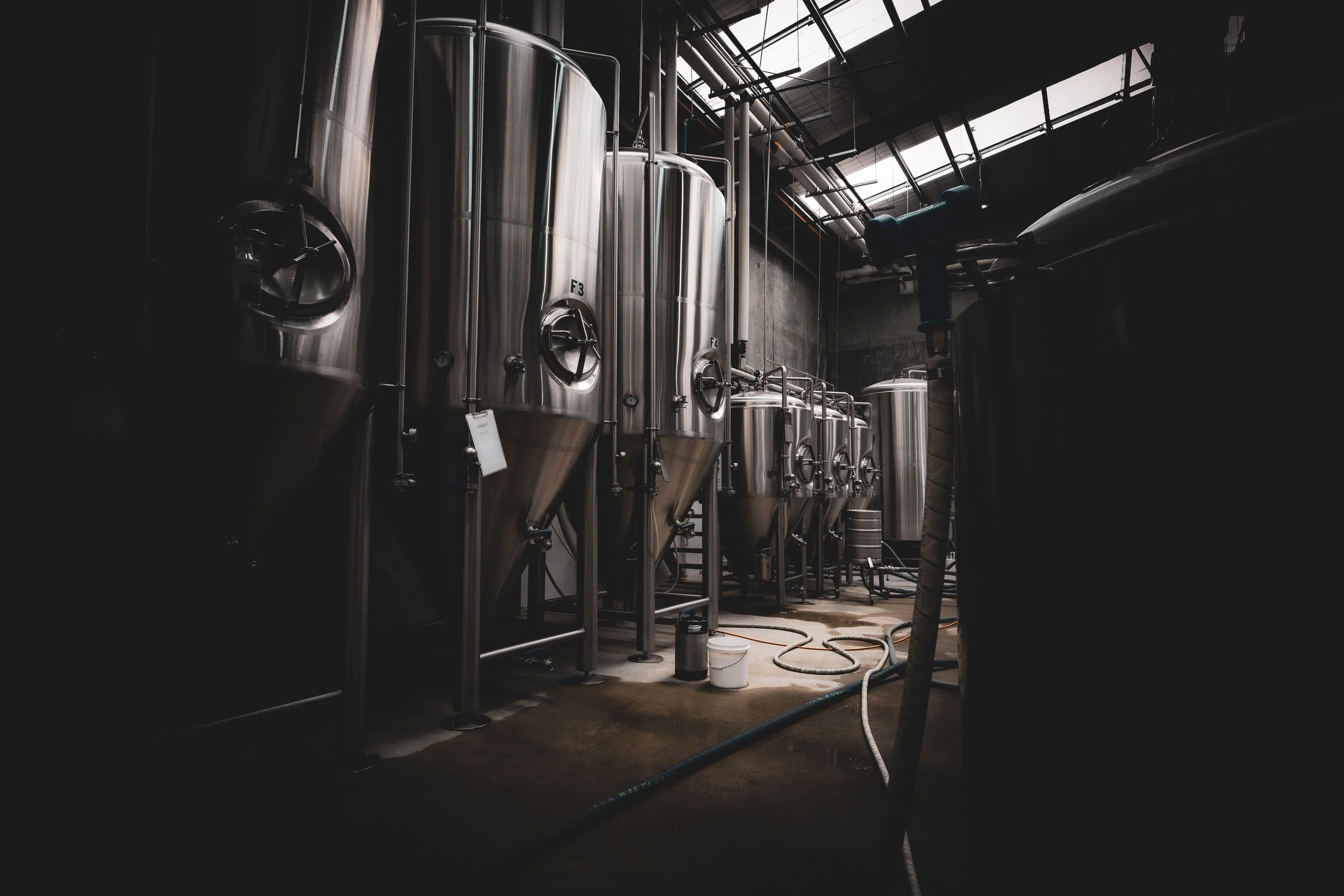 Large stainless steel fermentation tanks in a brewery or industrial facility with pipes and hoses connected, and natural light coming through overhead windows.