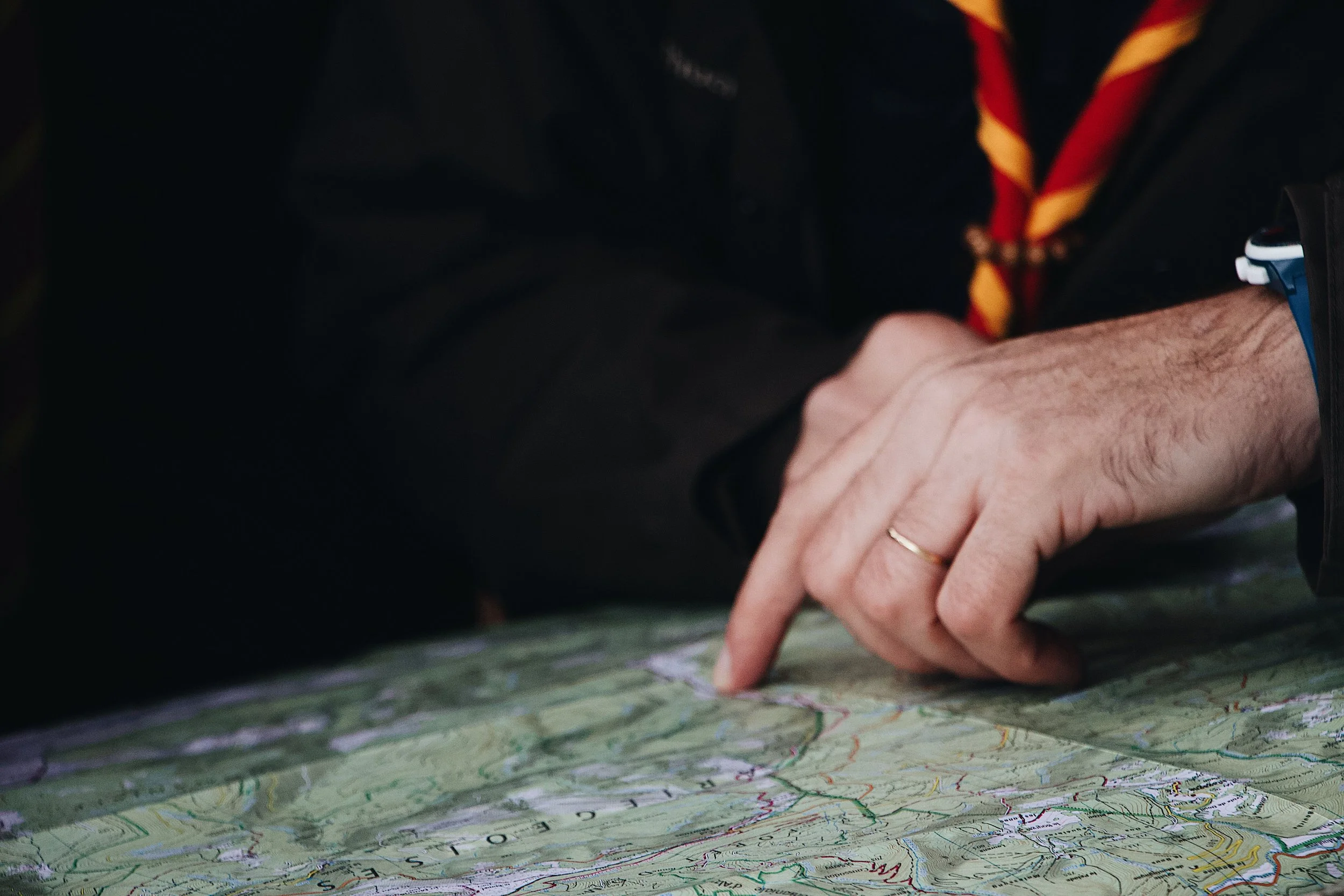 A person, likely a scout, is kneeling on the ground and pointing at a colorful map with various lines and markings. They are wearing a black jacket with a red and yellow striped neckerchief and a blue watch on their wrist.