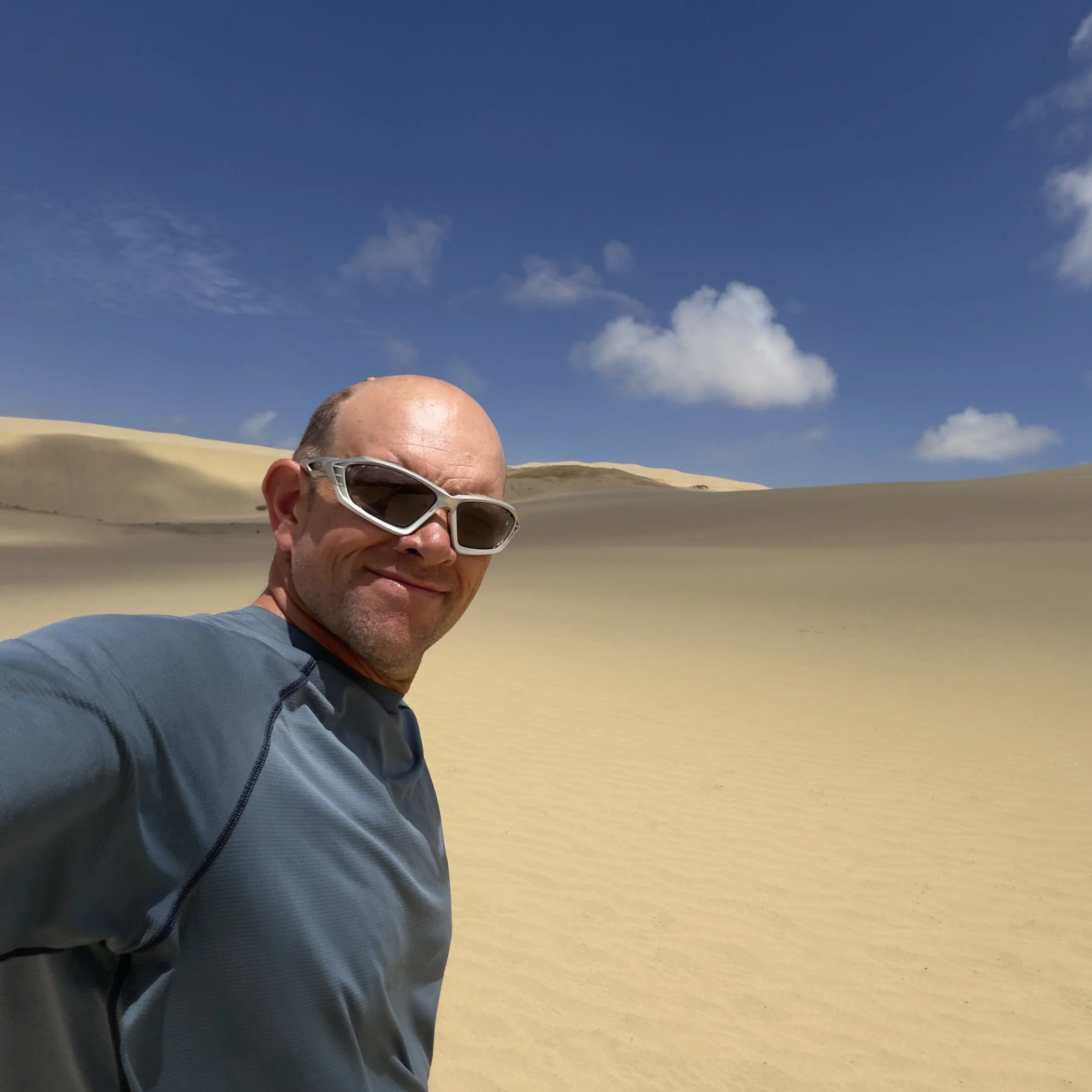 Close-up of a person smiling wearing sunglasses and a blue jacket, with sand dunes and a blue sky with scattered clouds in the background.