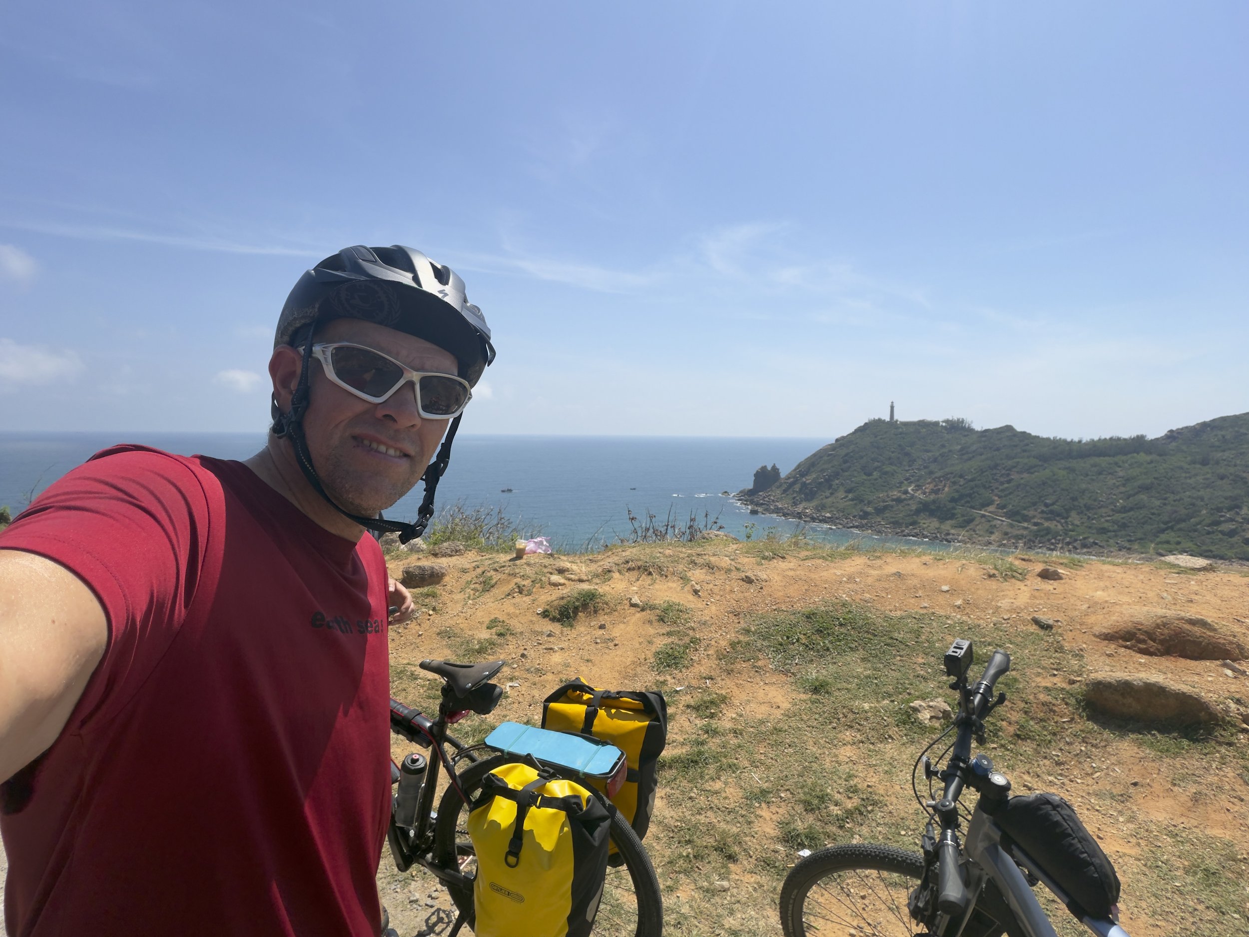 A man wearing a black helmet and white sunglasses taking a selfie on a mountain trail, with bicycles and a coastal landscape featuring a lighthouse in the background.