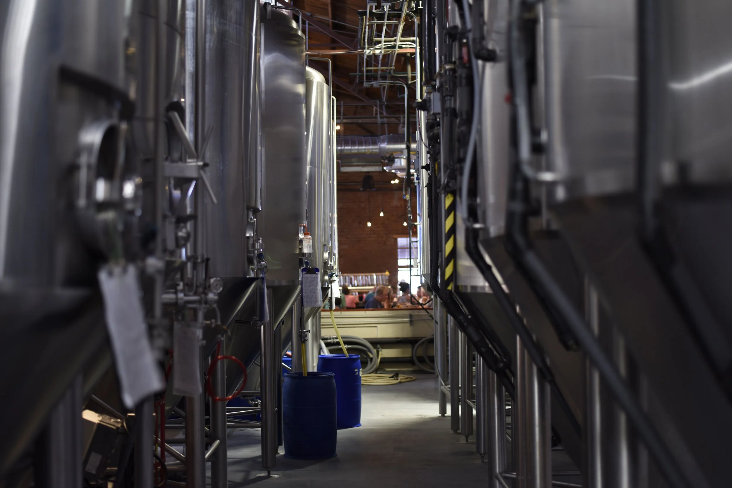 View of large stainless steel brewing tanks with pipes and hoses in a brewery, with some people working and sitting at tables in the background.