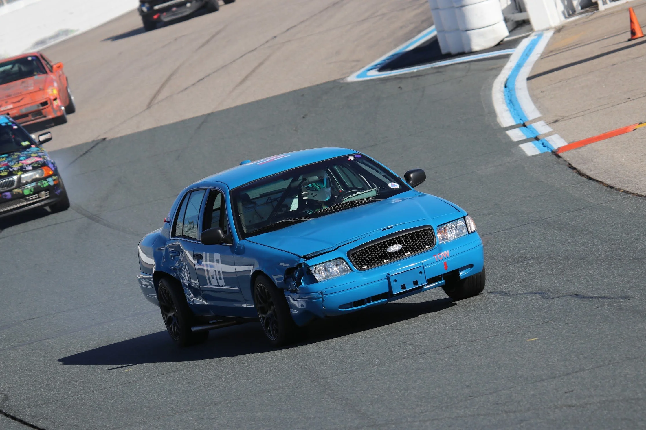 A blue car involved in a racing accident, with damage to the front and side, on a race track with other cars in the background.