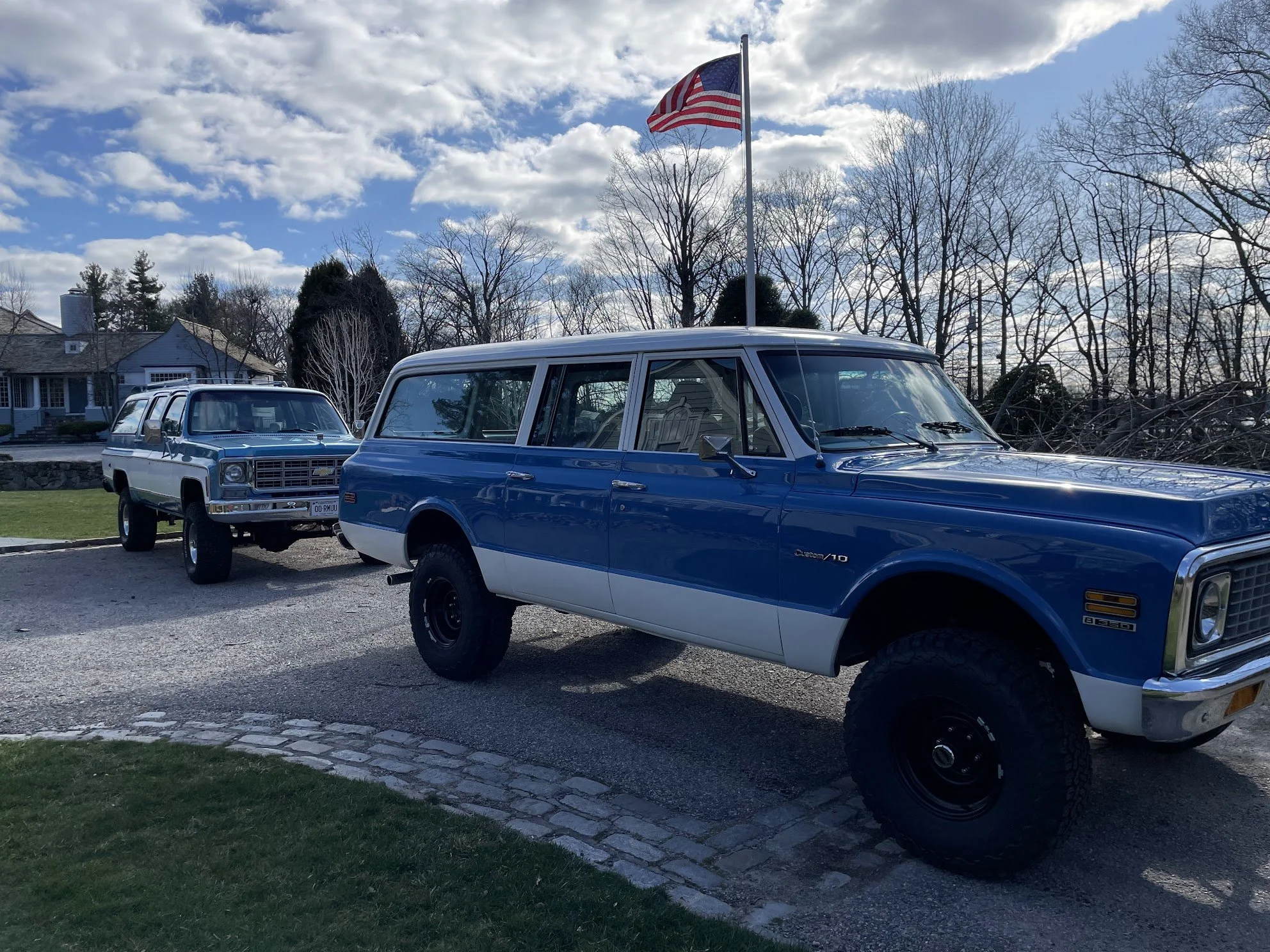 Two vintage SUVs, one blue with a white bottom, parked outdoors on a gravel driveway under a partly cloudy sky, with an American flag on a pole behind them and trees in the background.