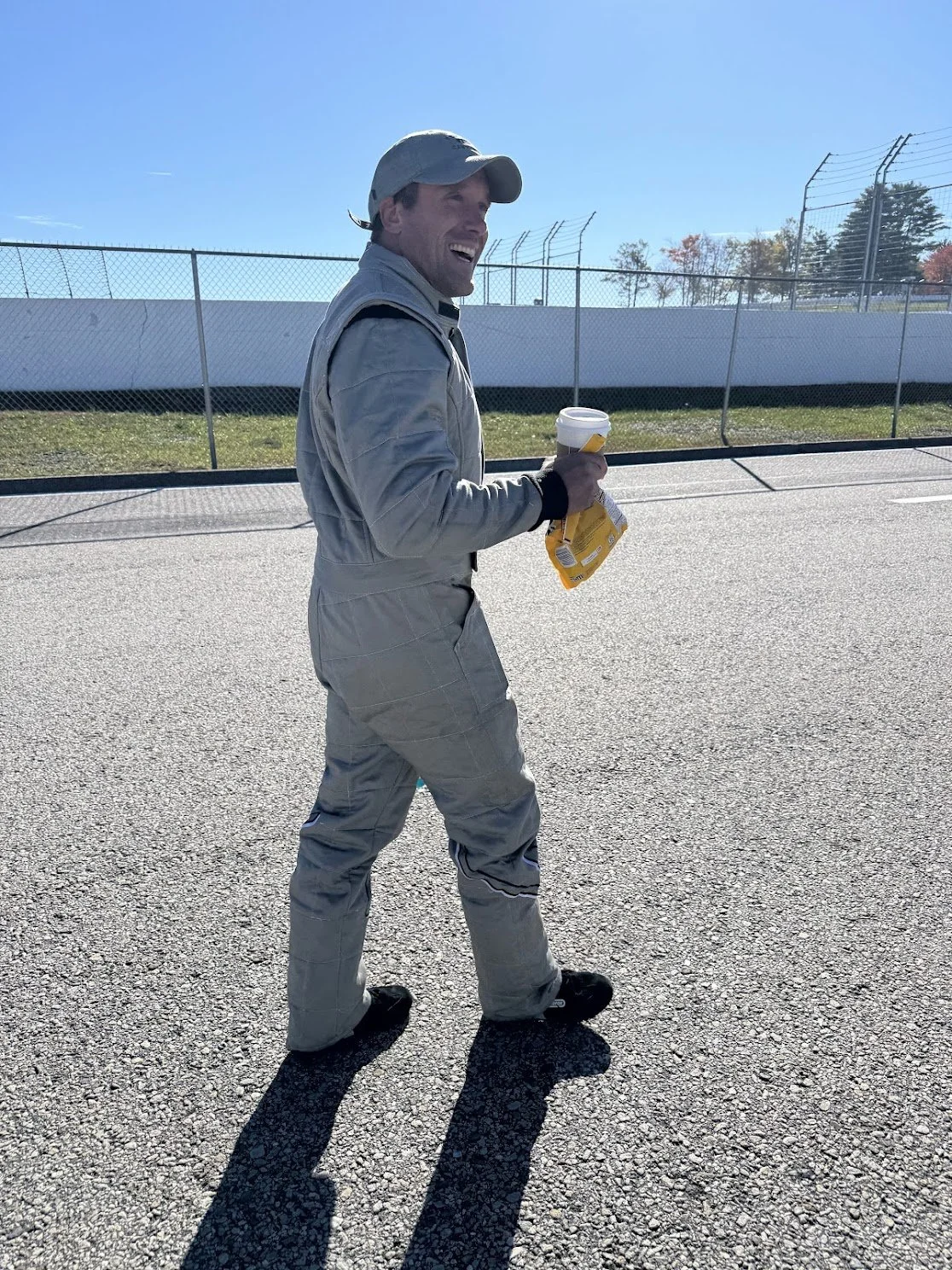 A man in racing suit smiling and walking on a racetrack, holding a coffee cup and a snack.