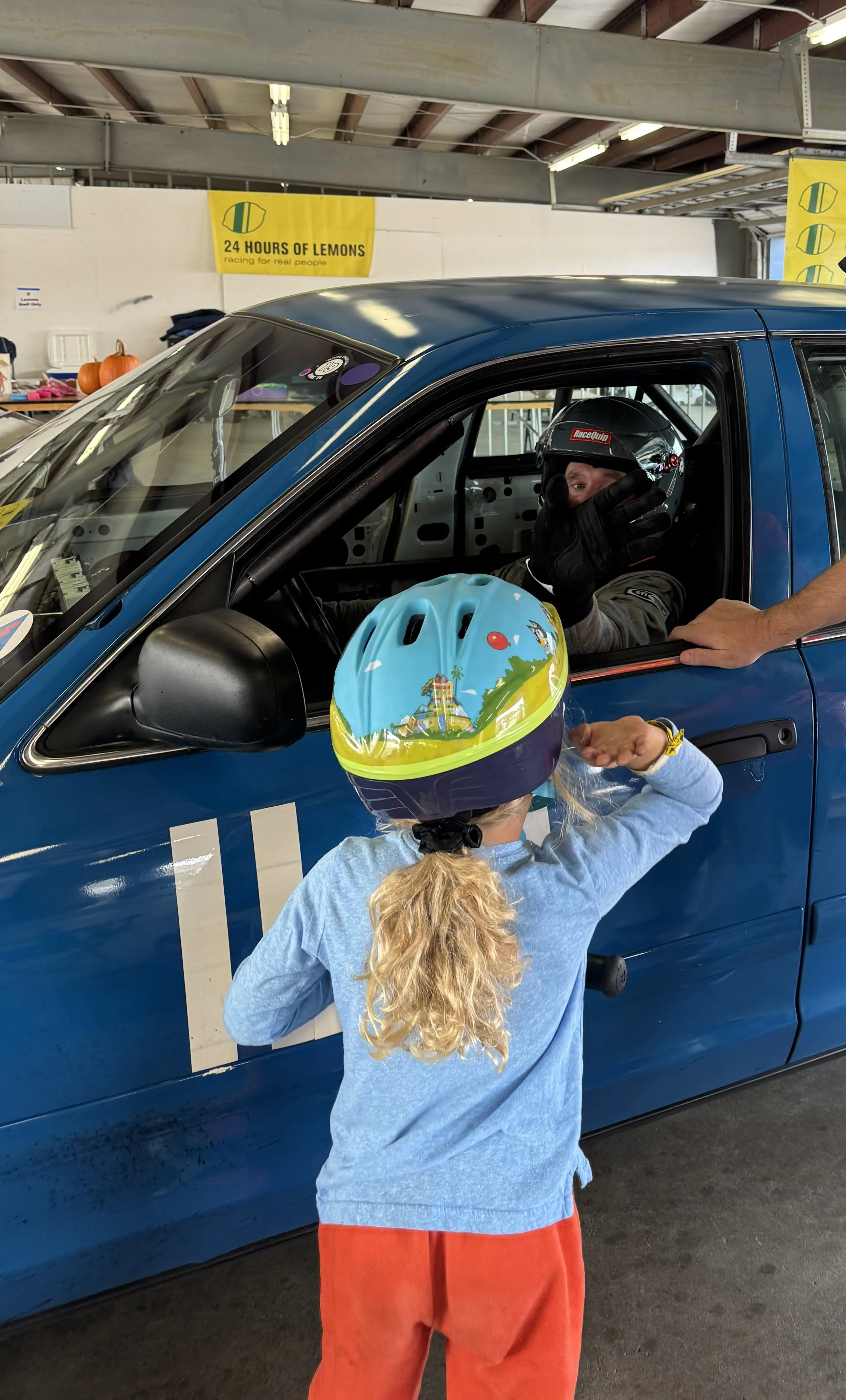 A young girl wearing a colorful bicycle helmet and red pants, talking to a race car driver inside a blue race car with a helmet and racing gear. The scene is inside an indoor racing facility with a sign in the background that reads '24 Hours of Lemons'.