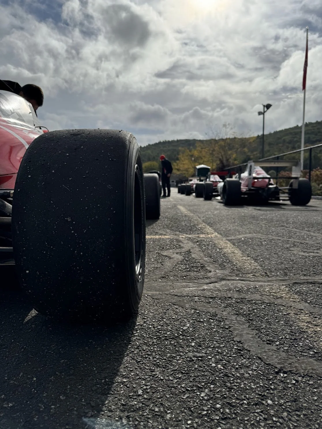 Close-up of a racing car tire on tarmac with other race cars and people in the background, under a partly cloudy sky.