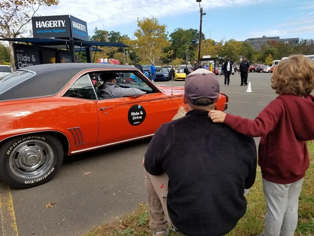 An orange vintage car with a black roof and a 'Ride & Drive' sign on its door, parked in a lot with other vehicles. Two children, one sitting and one standing with an arm around the other, watch the car. Several people are seen in the background, some wearing masks, near a service area with a sign that says 'Hagerty'.