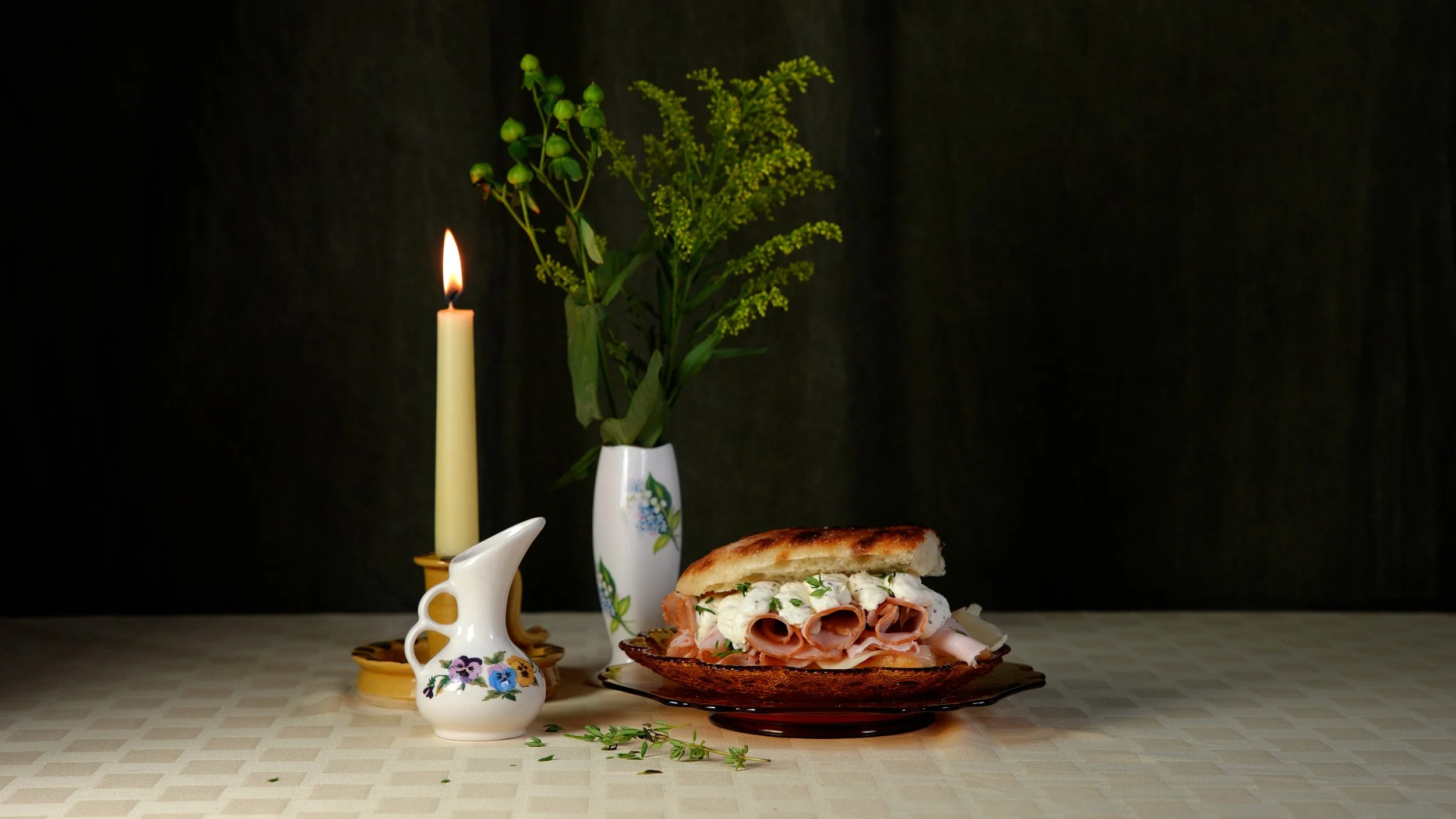A still life of a lit candle, a vase of green flowers, a small decorative pitcher with flowers, and a sandwich with meat and cheese on a dark plate, all on a beige checkered tablecloth against a dark background.