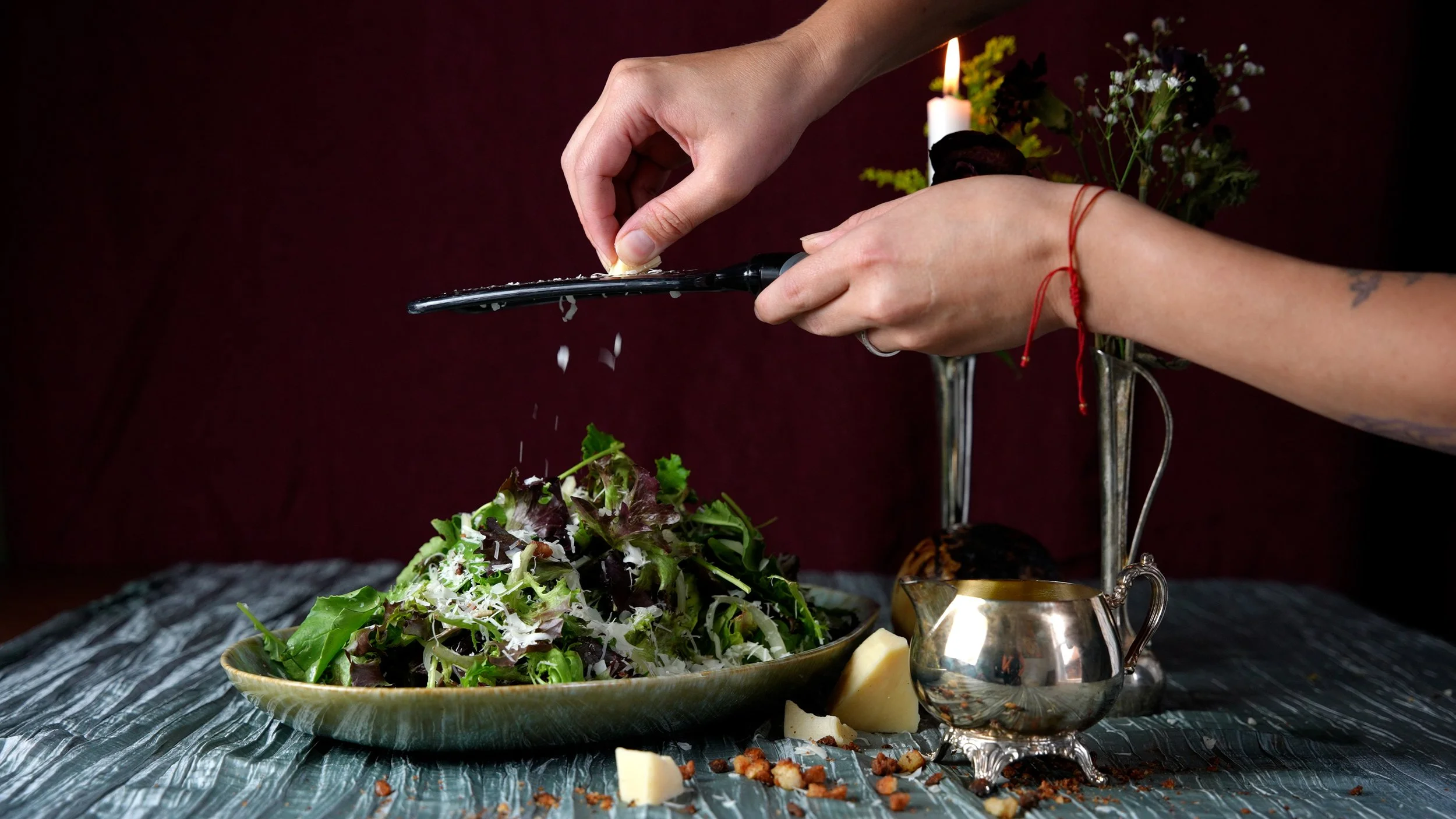 Hands sprinkling salt onto a fresh green salad at a table with cheese, nuts, and a silver gravy boat, with a lit candle and flowers in the background.