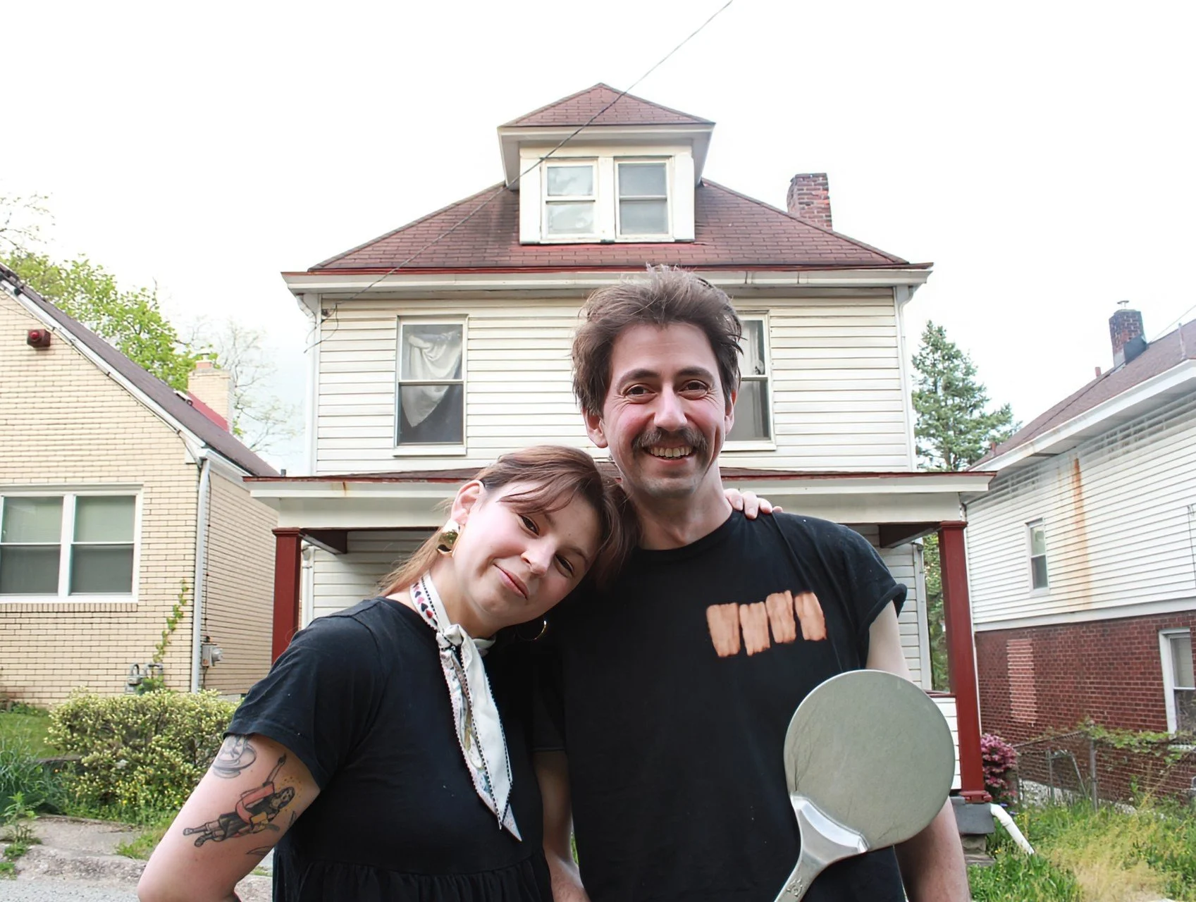 A smiling man and woman stand close together outside in front of a two-story house. The man holds a tennis racquet and wears a black t-shirt. The woman has long brown hair, a black top, and visible tattoos on her arm. The house has white siding, a red roof, and several windows.