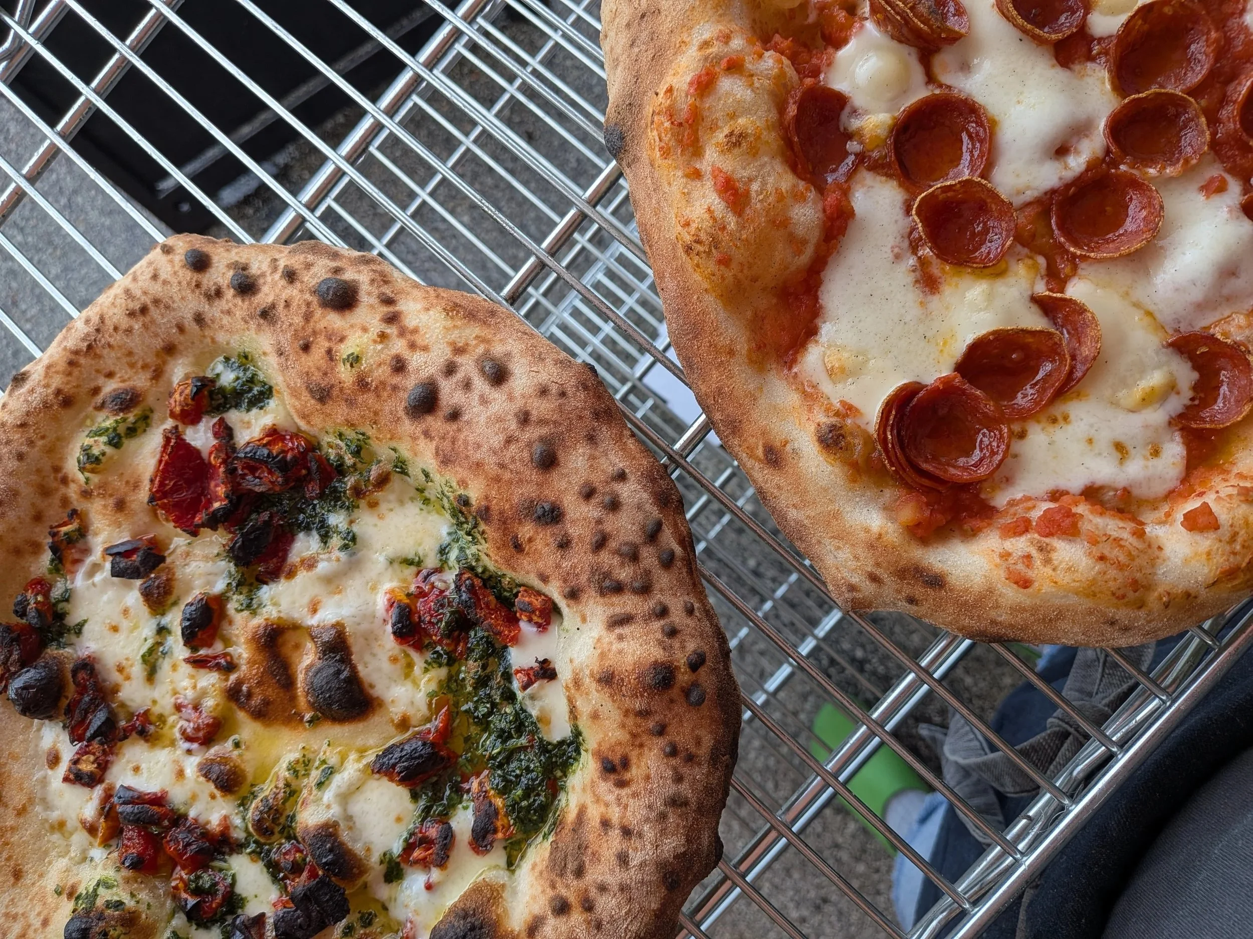 Close-up of two different personal-sized pizzas on a metal cooling rack. One has cheese and pepperoni, the other has cheese, herbs, and chili peppers.