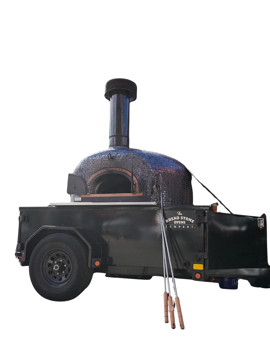 A mobile outdoor pizza oven setup on a black trailer, with a domed oven covered in small dark tiles, a chimney at the top, and pizza peels leaning against the trailer side.