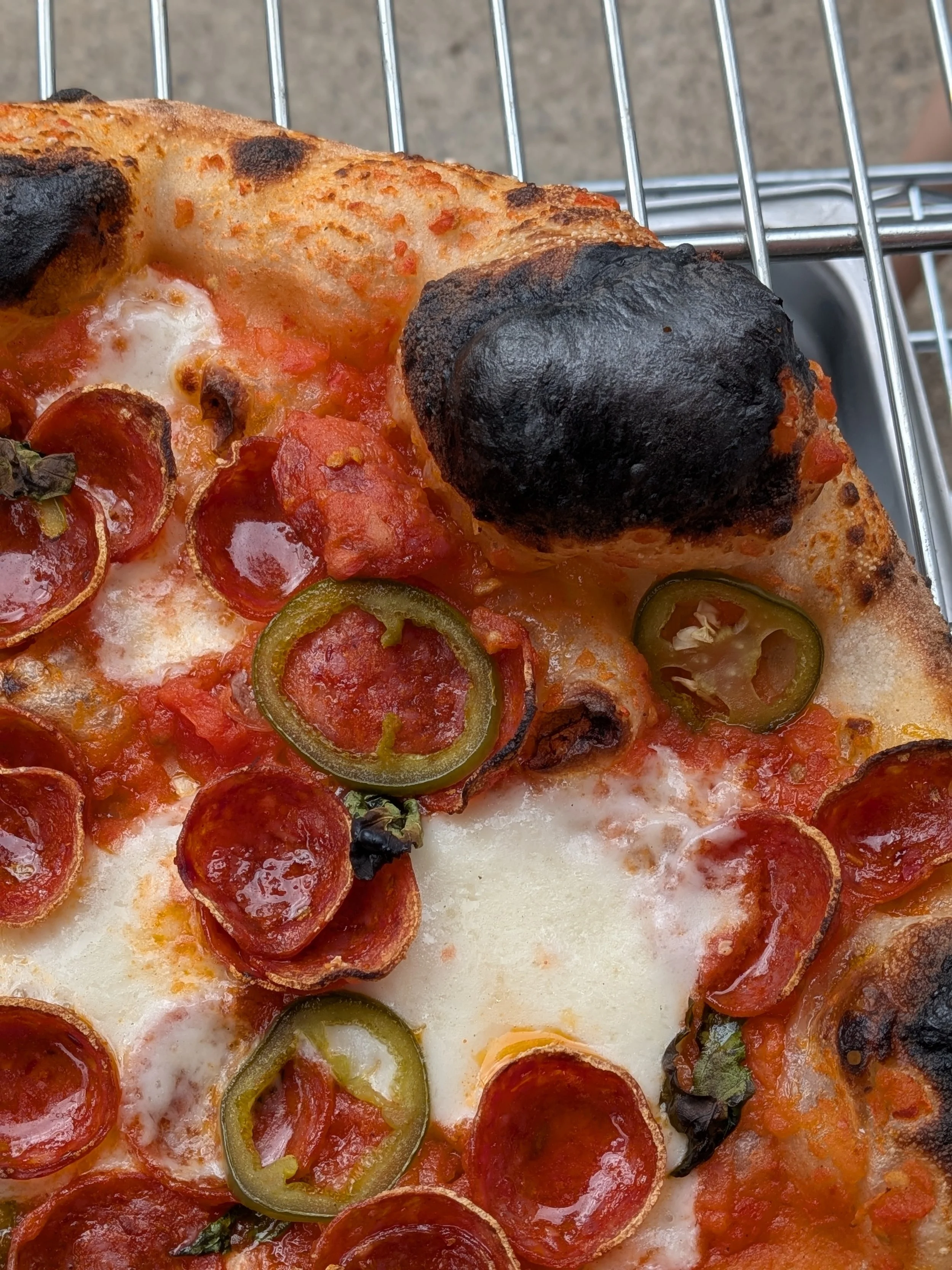 Close-up of a pepperoni pizza with jalapeño slices, some charred crust spots, and a blackened spot on the crust, on a wire rack