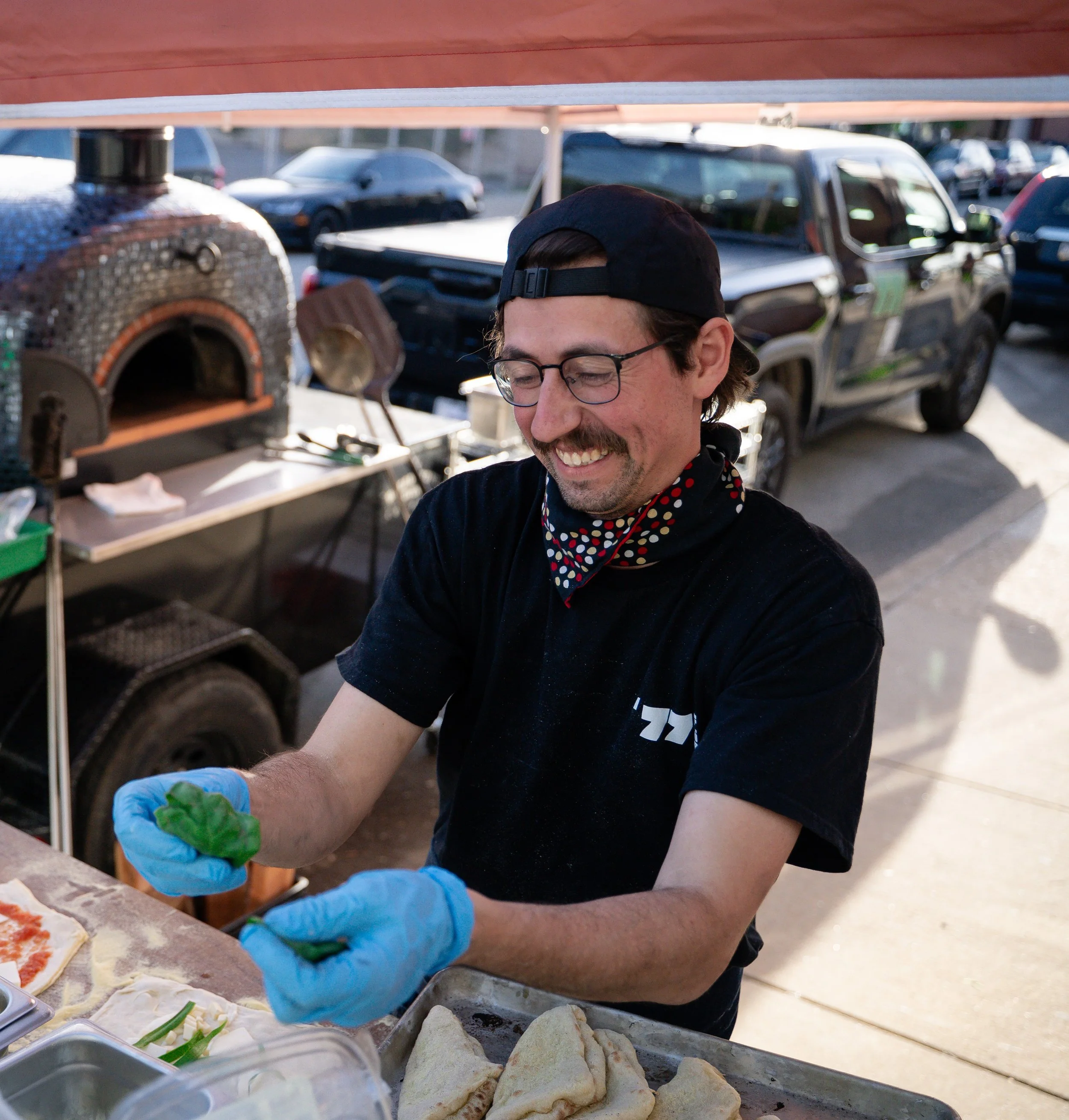 A man smiling and preparing food at an outdoor food stand, wearing gloves, a black shirt, and a face mask around his neck, with cars and a pizza oven in the background.