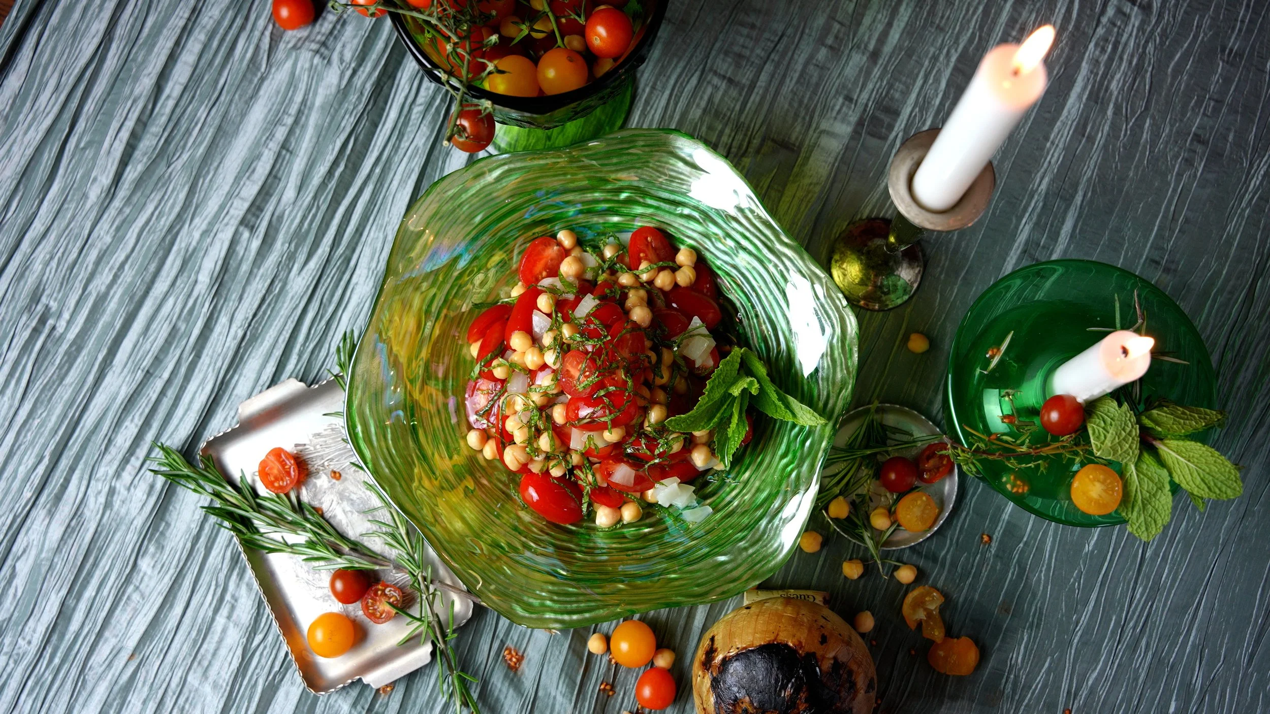 A colorful tomato salad in a green glass bowl on a rustic wooden table, with cherry tomatoes, fresh herbs, and candles nearby.