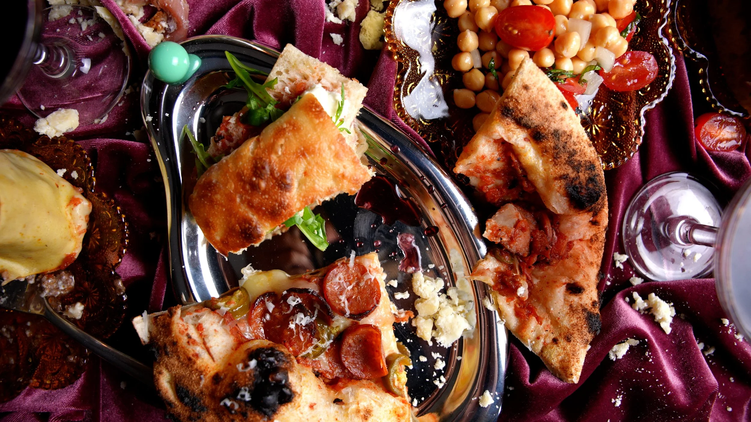 An assortment of pizza slices, chickpeas salad, and pastries on a decorative table setting with a purple cloth.