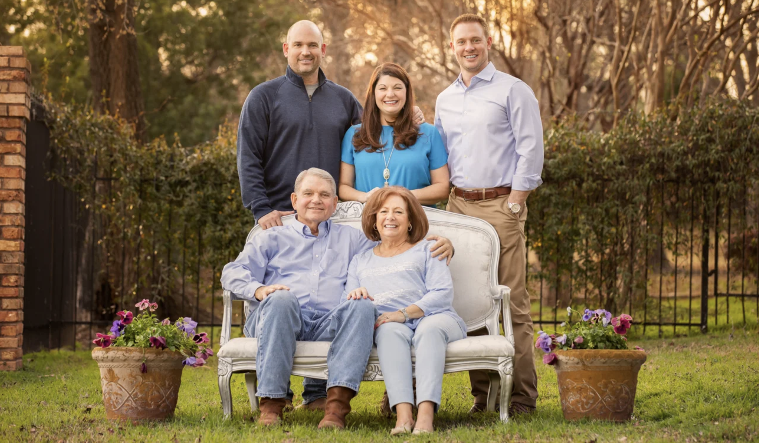A multigenerational family outdoors, with five adults, sitting and standing, smiling, with potted flowers in front, park-like fence and trees in the background.