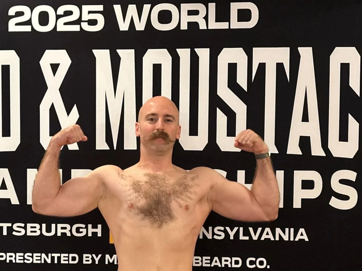 A shirtless man with a bald head, mustache, and chest hair flexing biceps in front of a black and white event banner.