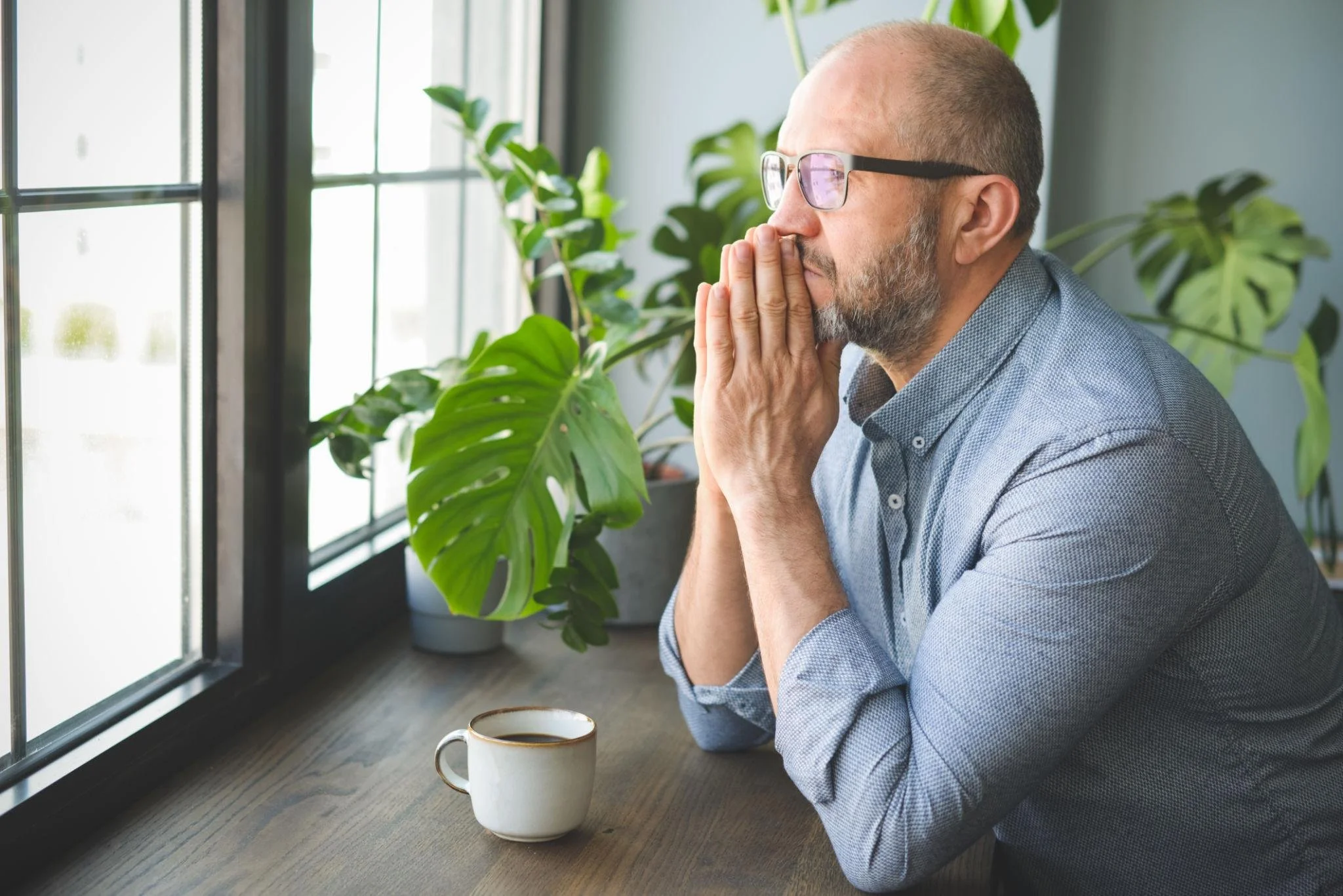Thoughtful man sitting at a table with clasped hands near his lips, looking out the window while processing stress and difficult emotions.