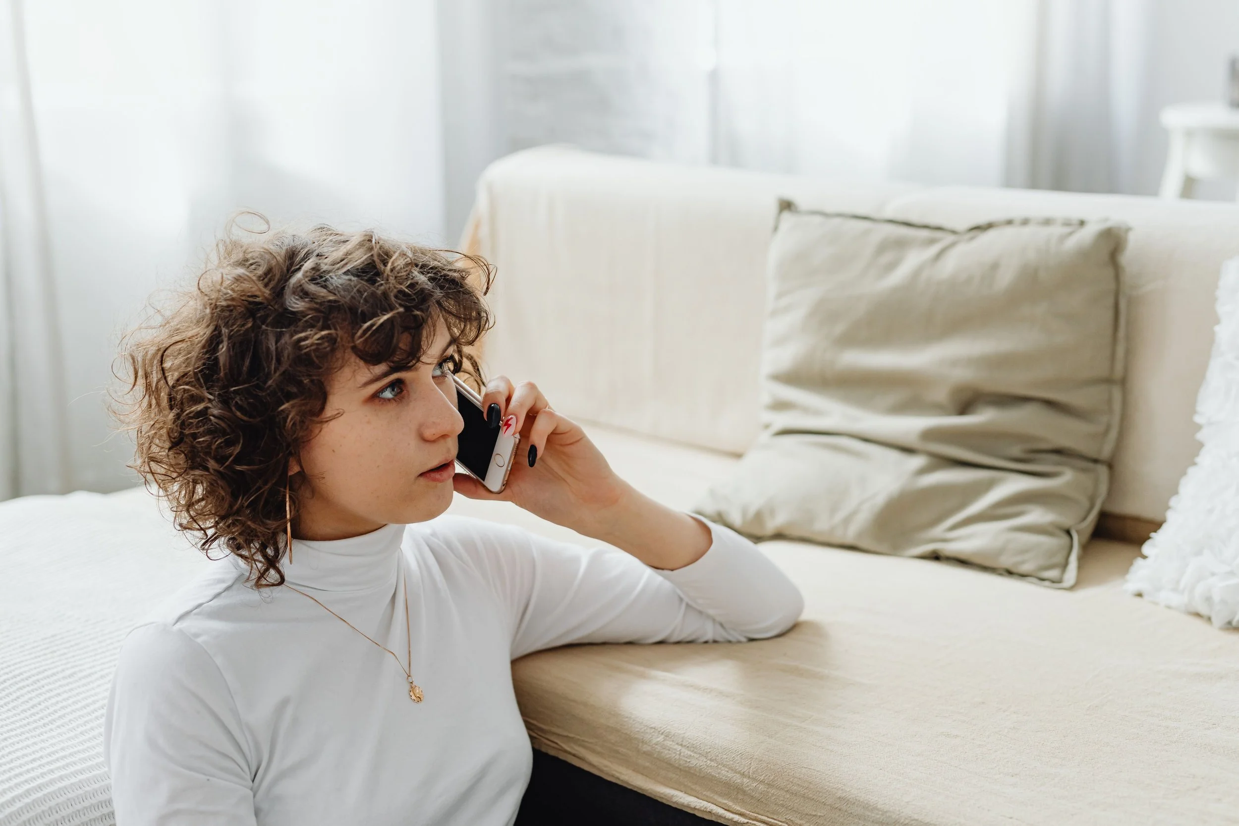Woman with curly hair sitting beside a couch, making a phone call for a consultation.