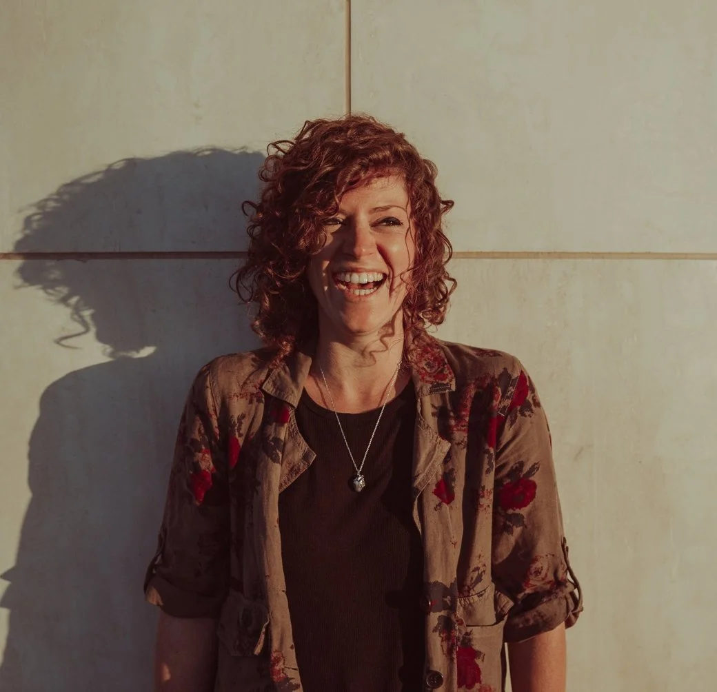 A woman with curly red hair, smiling, standing against a beige wall with her shadow cast on it, wearing a brown floral jacket and a black shirt.