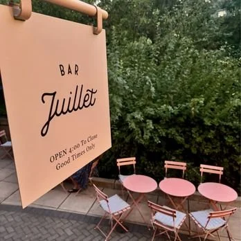 Outdoor seating area with pink chairs and tables next to a sign that reads 'Bar Juillet, open 4:00 to close, good times only'