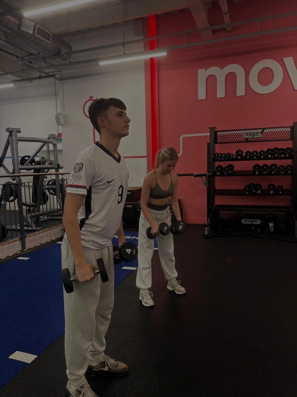 A man and woman lifting dumbbells in a gym. The man is wearing a white sports jersey with the number 9. The woman is wearing a dark sports bra and white workout pants. They are standing on a black and blue gym floor, with exercise equipment and a red wall with the word 'move' in the background.